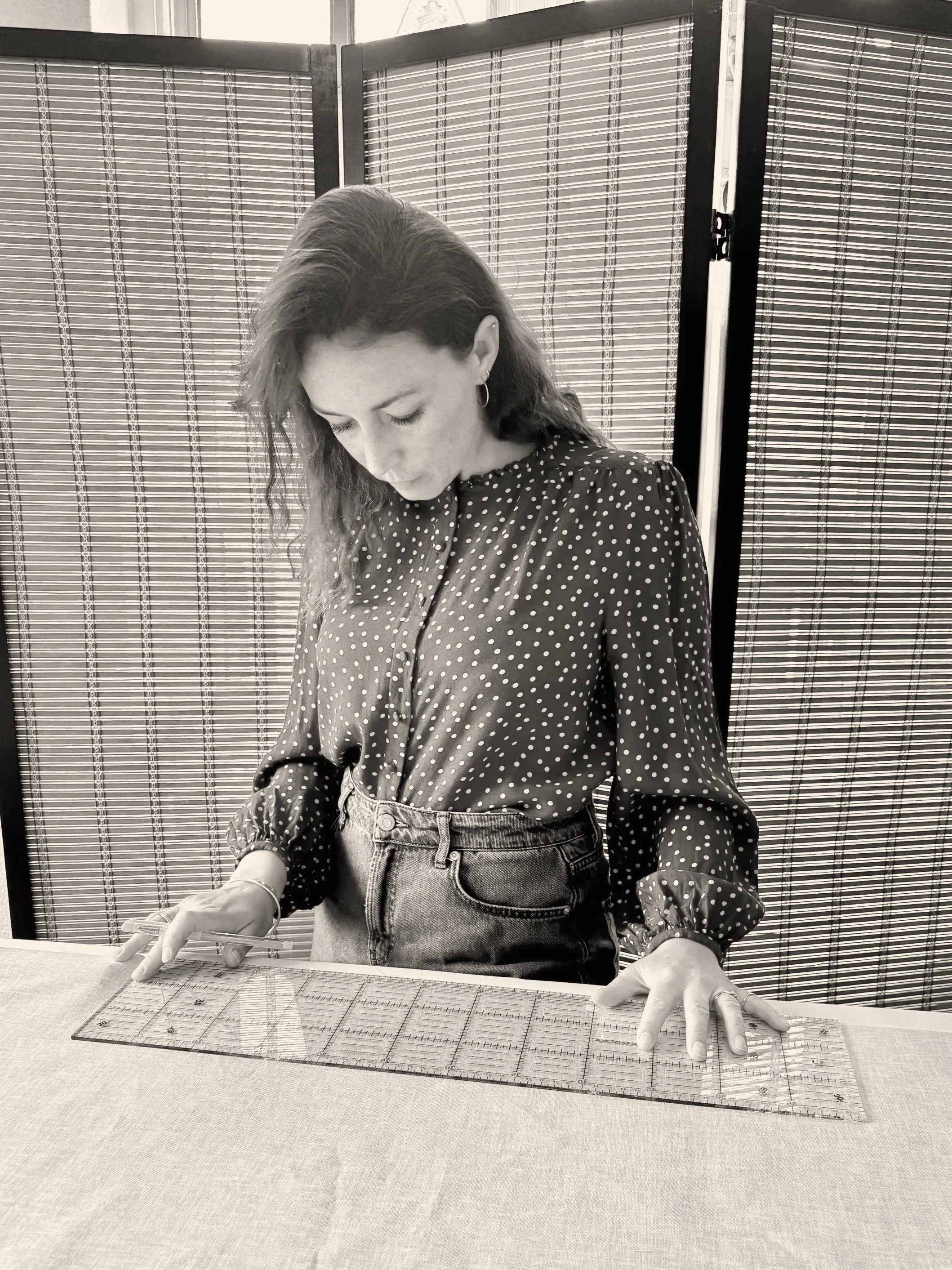 A woman with wavy hair wearing a polka dot blouse and jeans is standing at a table with a large cutting mat, holding a pencil in her right hand and her left hand resting on the table. She is looking down at her work, and behind her is a folding partition with patterned panels.