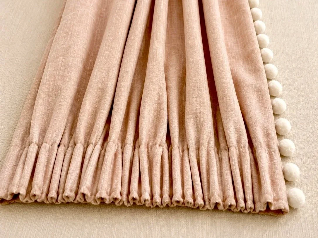 Light pink velvet curtain with white pom-pom trim, folded neatly on a beige surface.