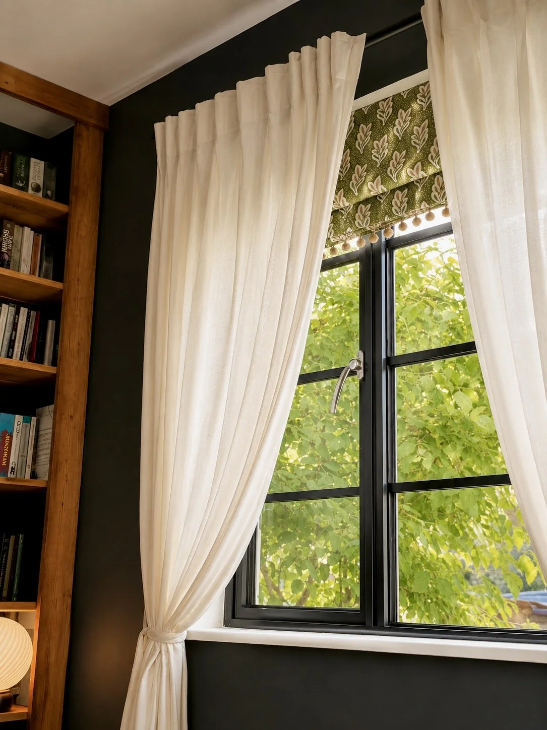 A window with white curtains partially drawn and a patterned valance with green and white leaf design. Outside visible green leaves on trees.
