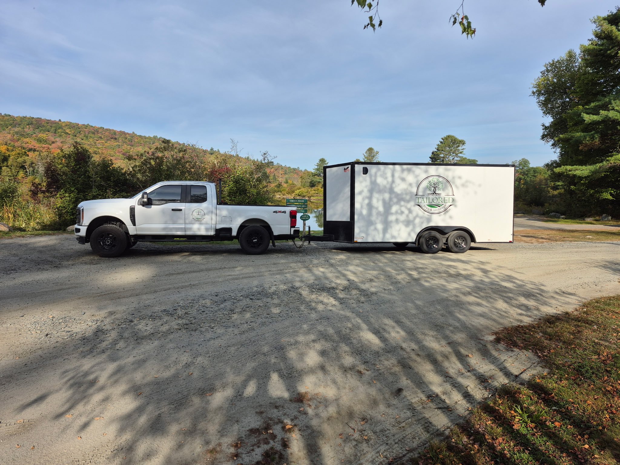 A white pickup truck towing a white enclosed trailer with a logo reading 'Tailored' parked on a gravel area near trees and a scenic landscape of hills with autumn foliage.