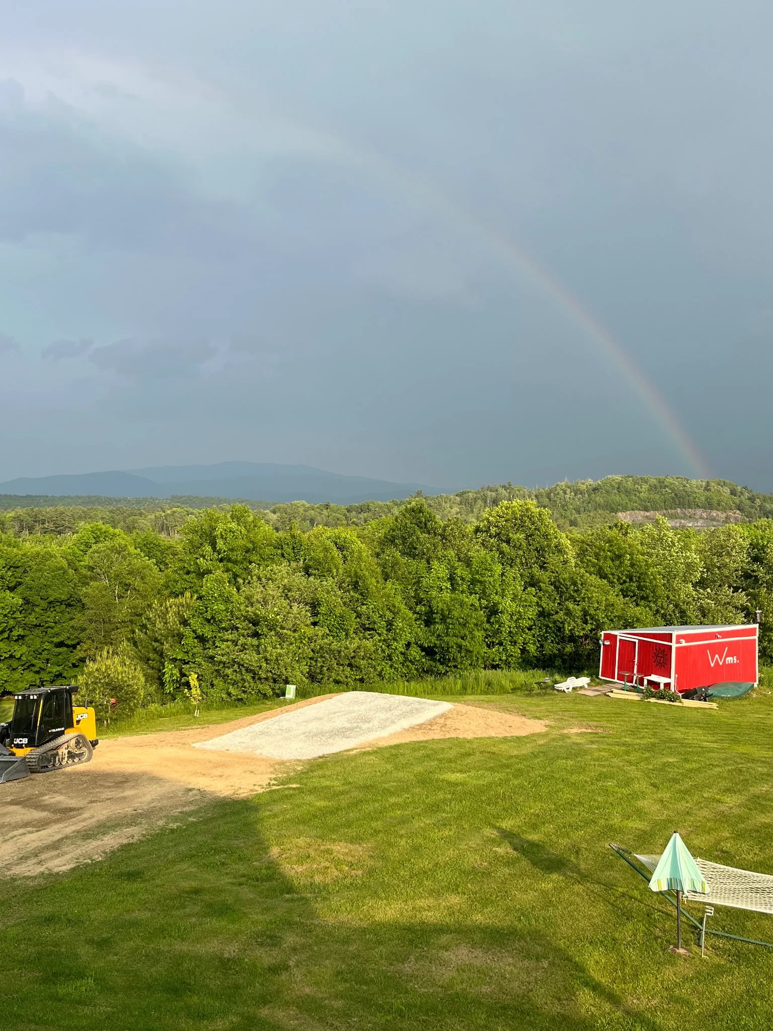 A landscape with a rainbow in the sky over green trees, a red shed with white trim, a hammock with an umbrella, a construction vehicle, and a gravel area on a grassy field.