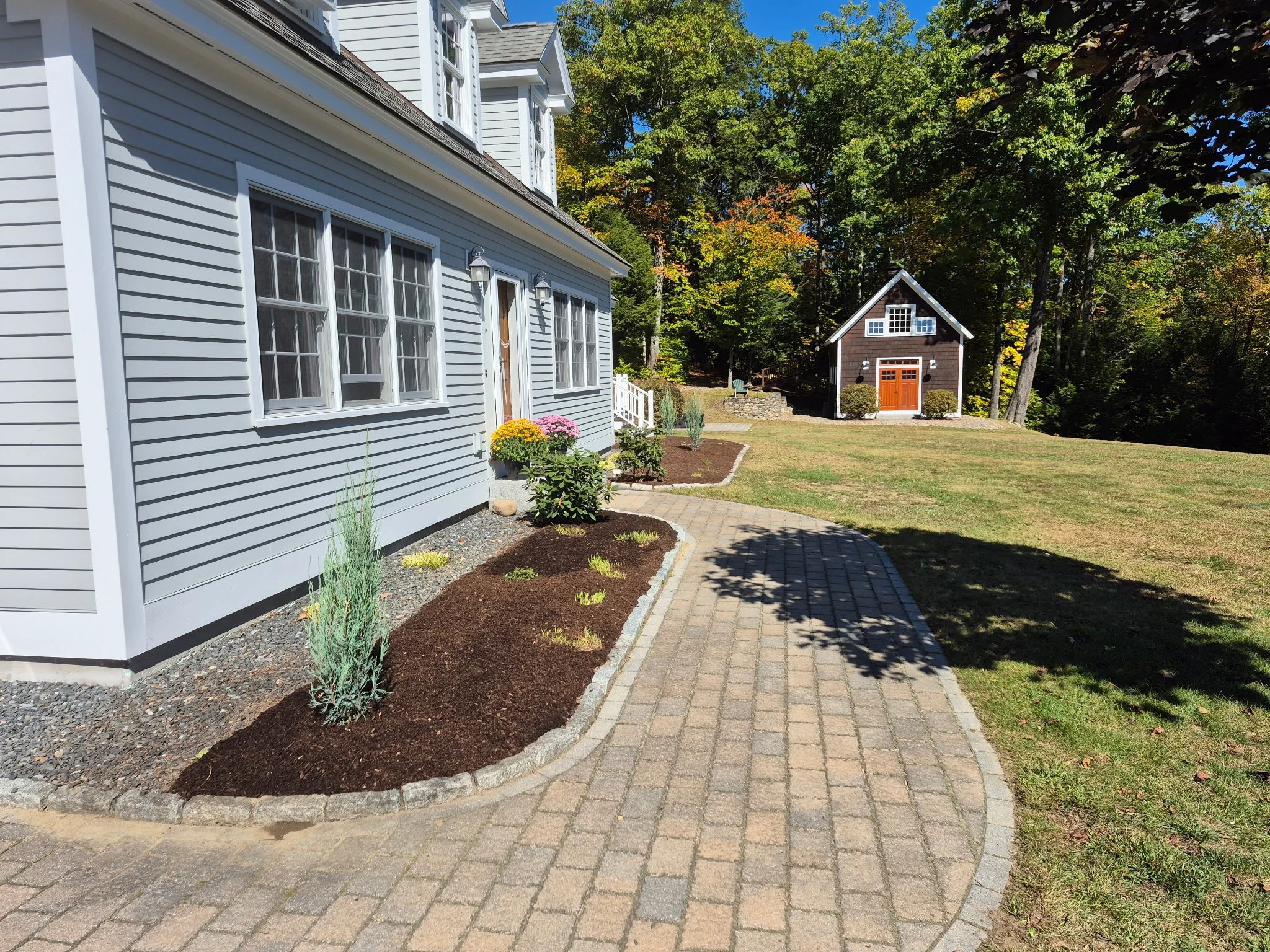A paved walkway runs past a gray house with white trim and multiple windows, with landscaped planting beds along the side. In the background, there is a small dark-colored barn or shed with a brown door, surrounded by a grassy yard and wooded area.
