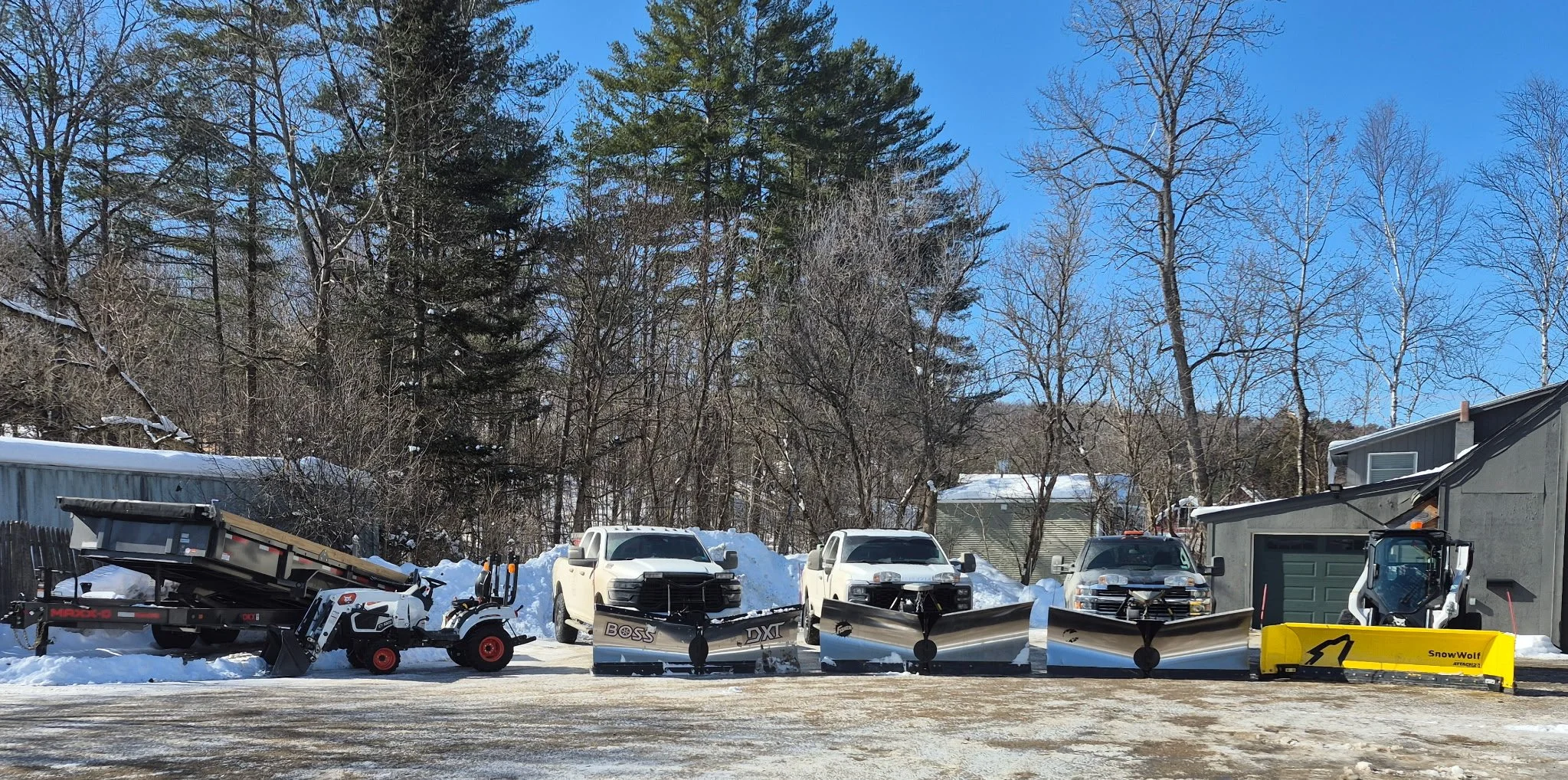 Snow removal equipment including snow plows and a snow blower, parked outdoors on a driveway with a snowy yard and trees in the background.
