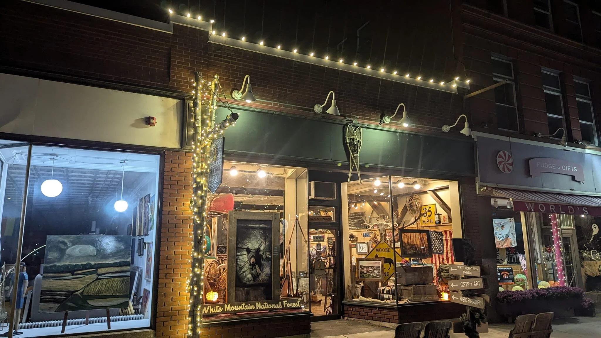 Night view of a storefront decorated with string lights and signs, filled with various artwork, furniture, and gift items inside, located in a small town.