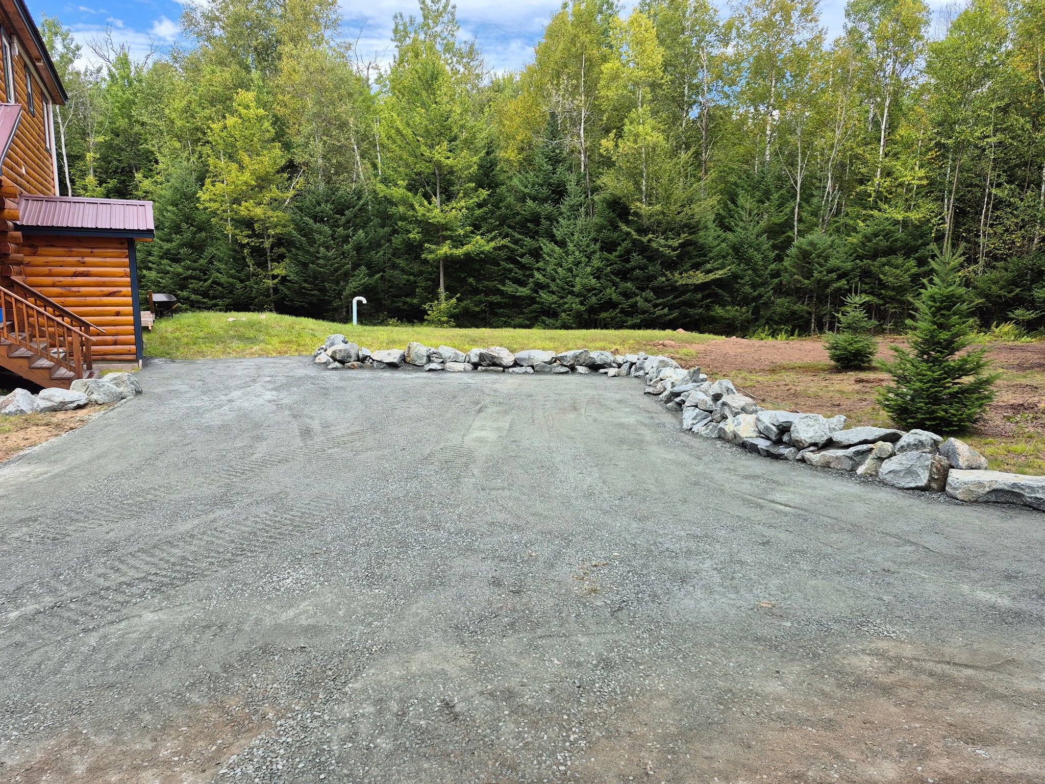 A gravel driveway with a line of large rocks along the edge, surrounded by green trees and a wooden house with stairs on the left, under a partly cloudy sky.