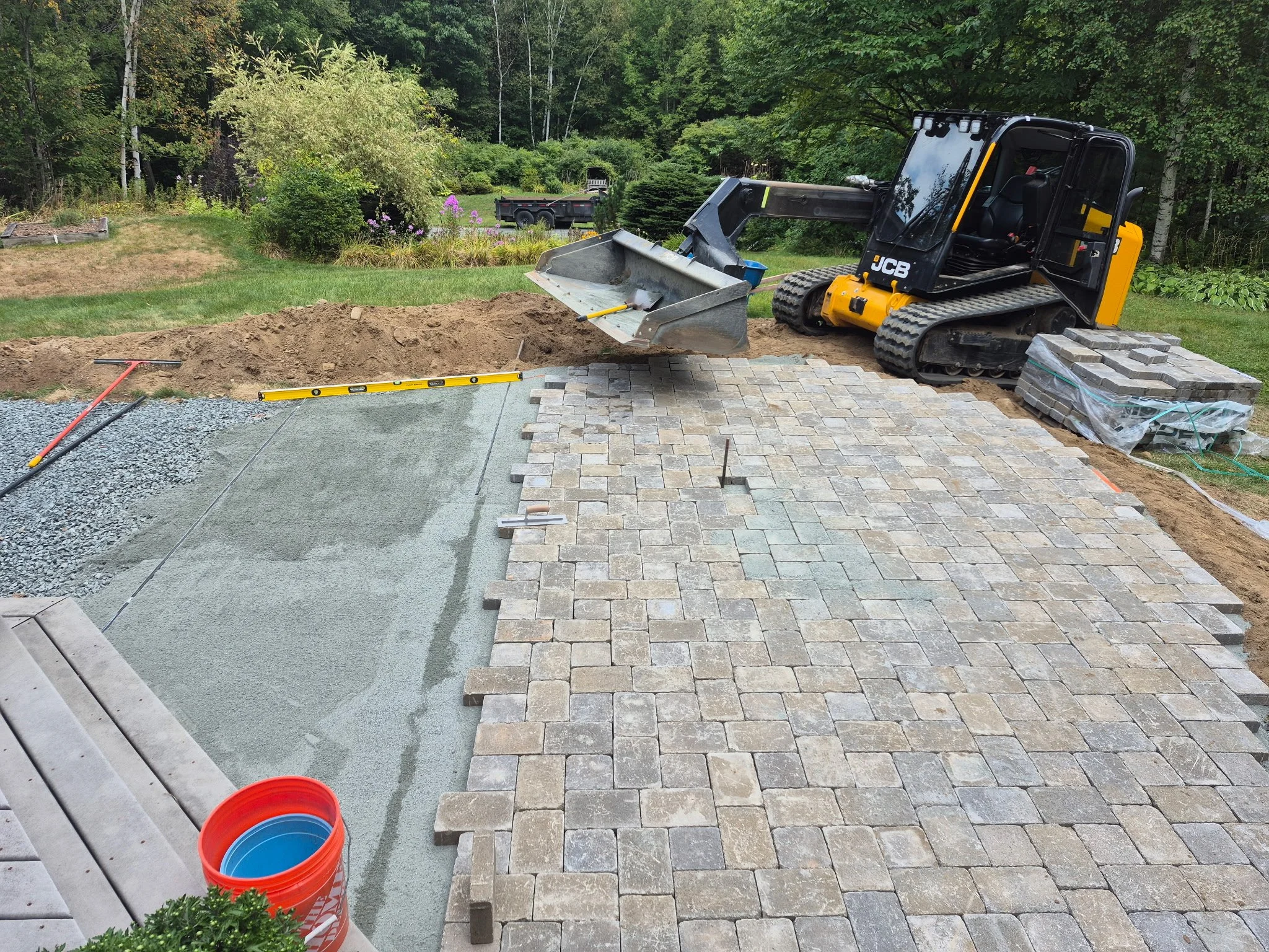 A small construction site shows a paver block patio being built. There is a small excavator with a bucket attachment on the right, and a level tool nearby. Pavers are being laid on a section of the patio, with some stacked on the side. A bucket with 