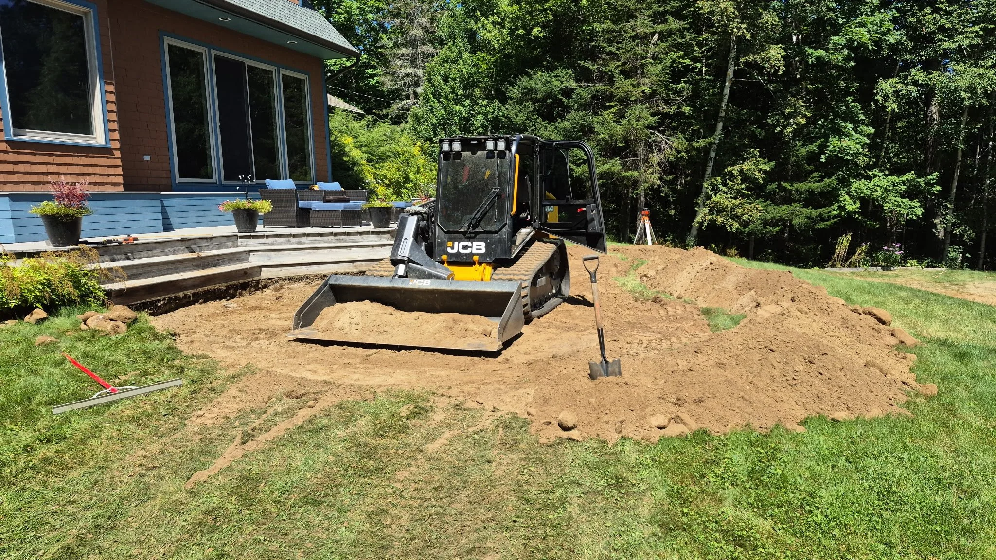 A small JCB skid steer loader working on a backyard yard project with a pile of dirt in front of it, near a house with a deck and potted plants, surrounded by trees and grass.
