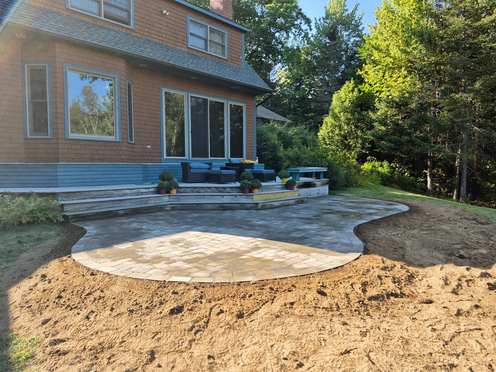 Backyard patio with partially completed paved stone surface, outdoor furniture, potted plants, and a house with large windows and sliding doors, surrounded by trees and greenery.