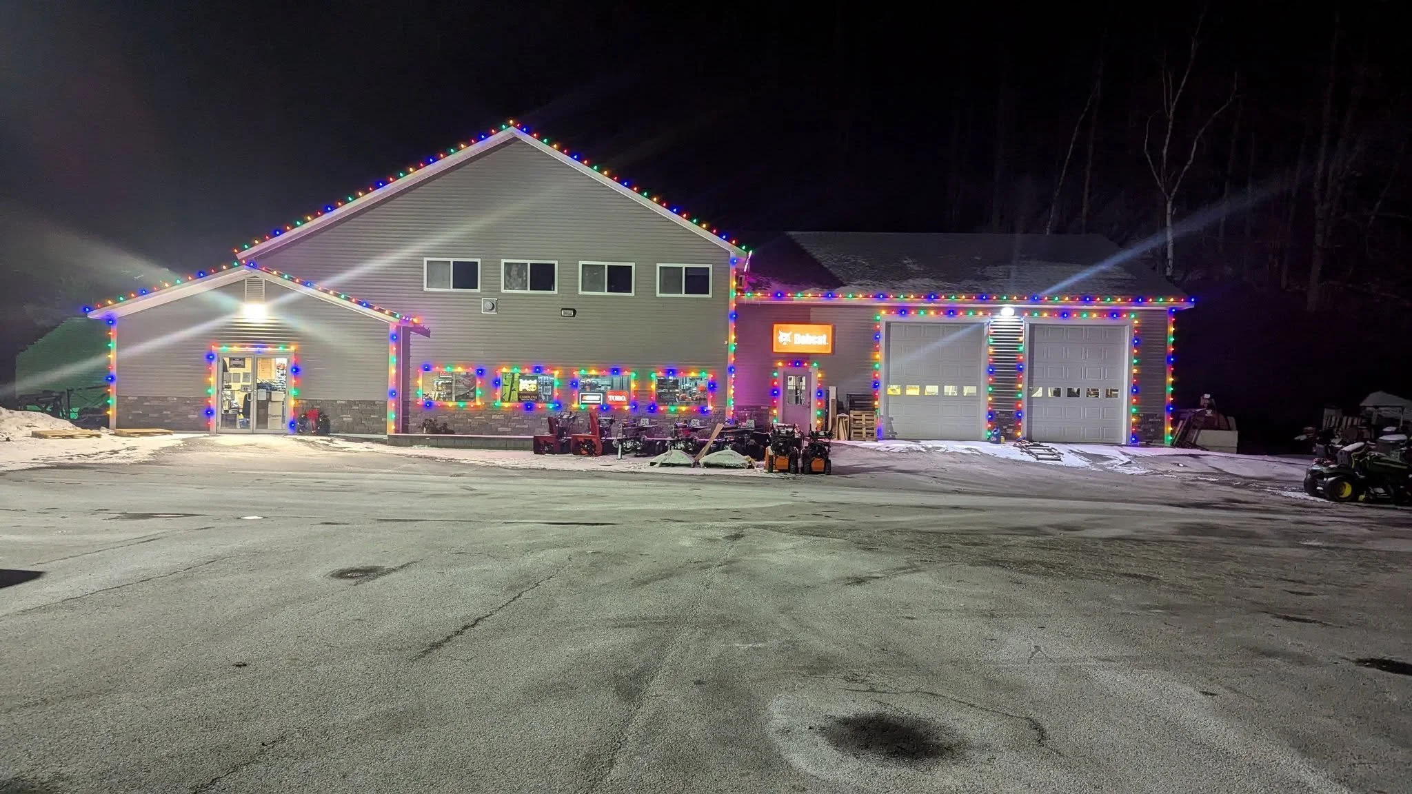 A building decorated with colorful Christmas lights at night, with snow on the ground and a parking lot in front.