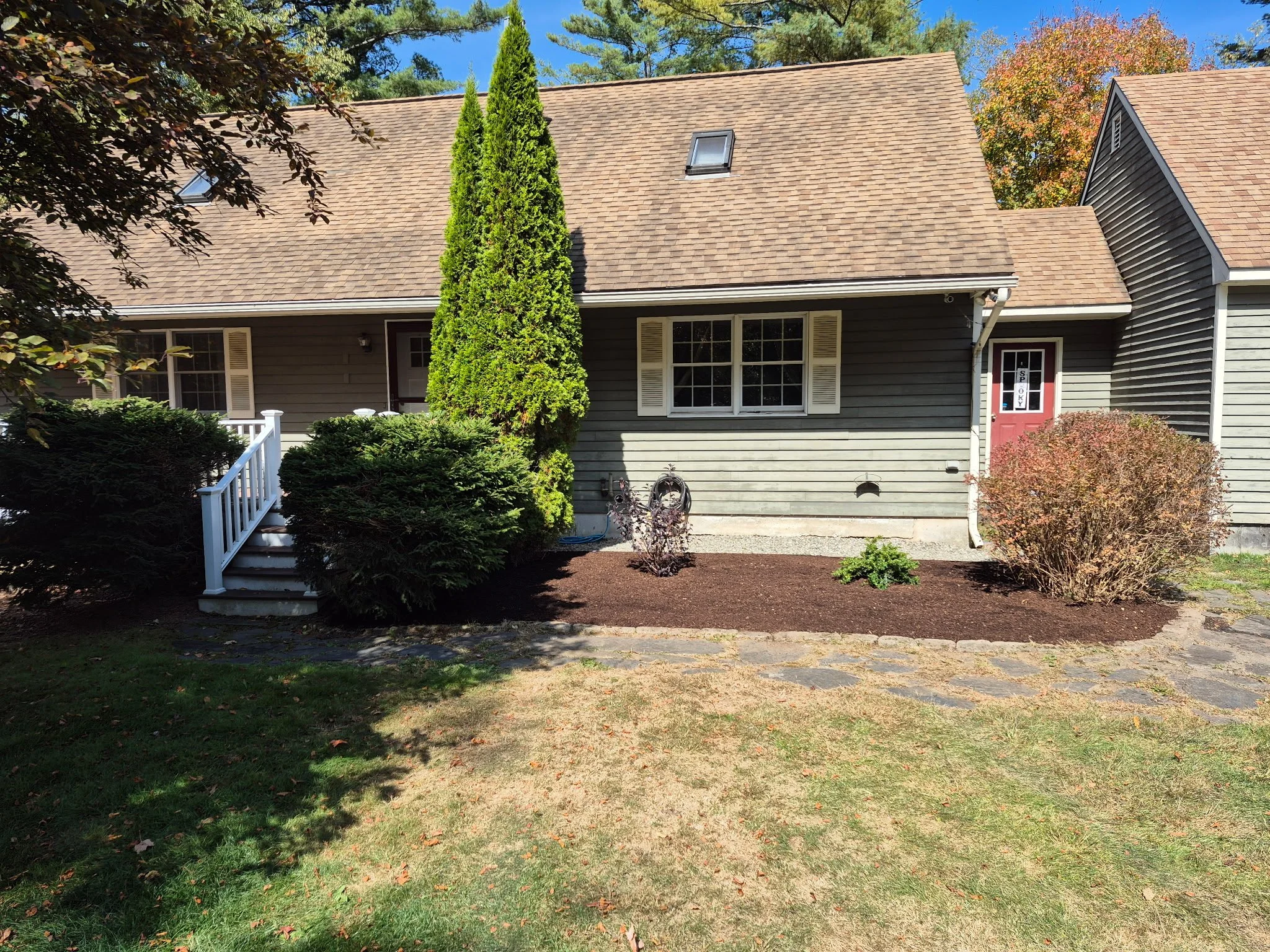 Front yard view of a house with gray siding, a pink front door with a decorative window, a small porch with white railing, and various shrubs and trees in front yard.