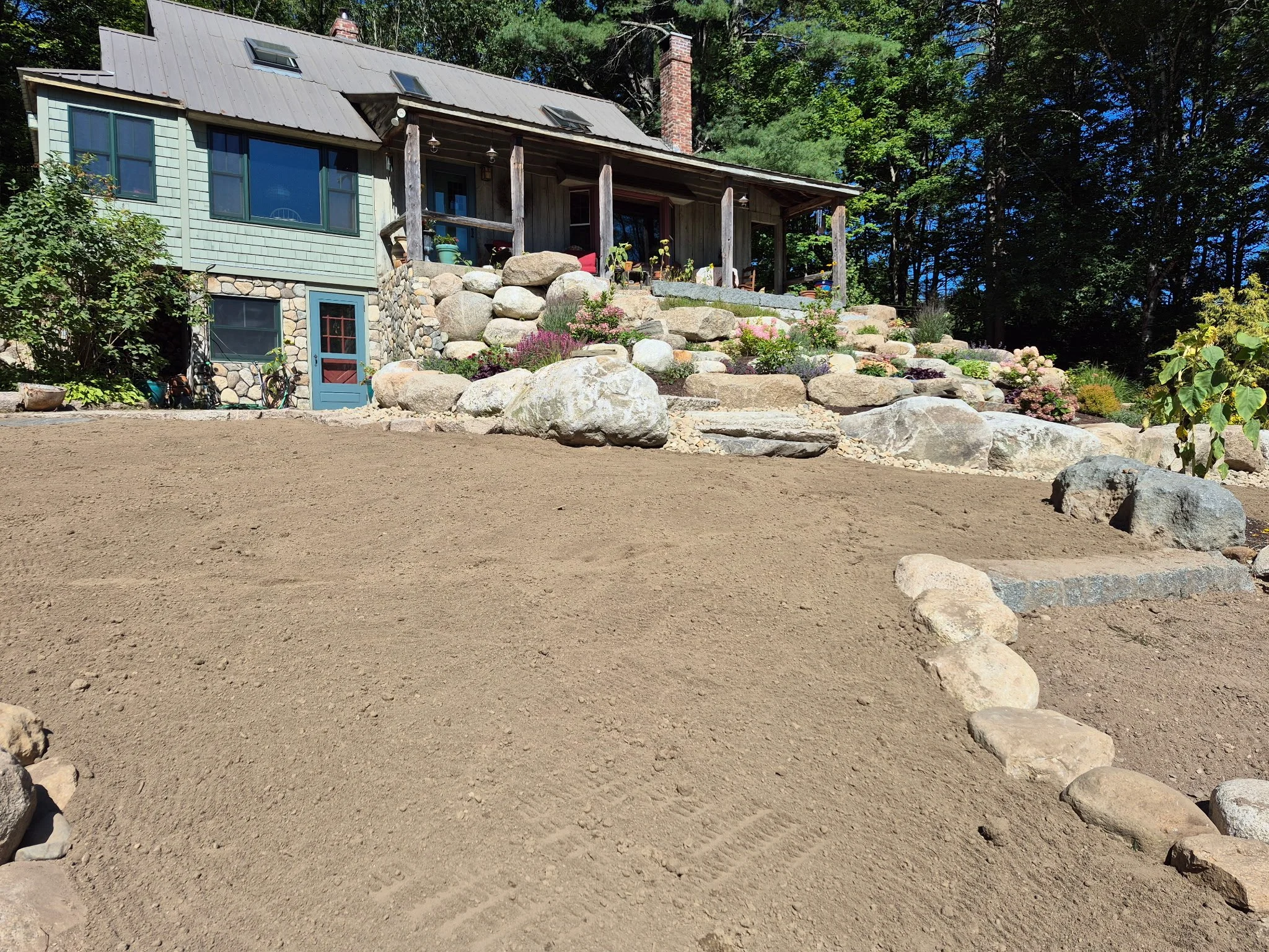 A house with a gray metal roof, green siding, and stone foundation, situated on a sloped yard. The yard features a large patch of tilled soil with a border of rocks, leading up to a garden with colorful plants and large rocks on a terraced slope.
