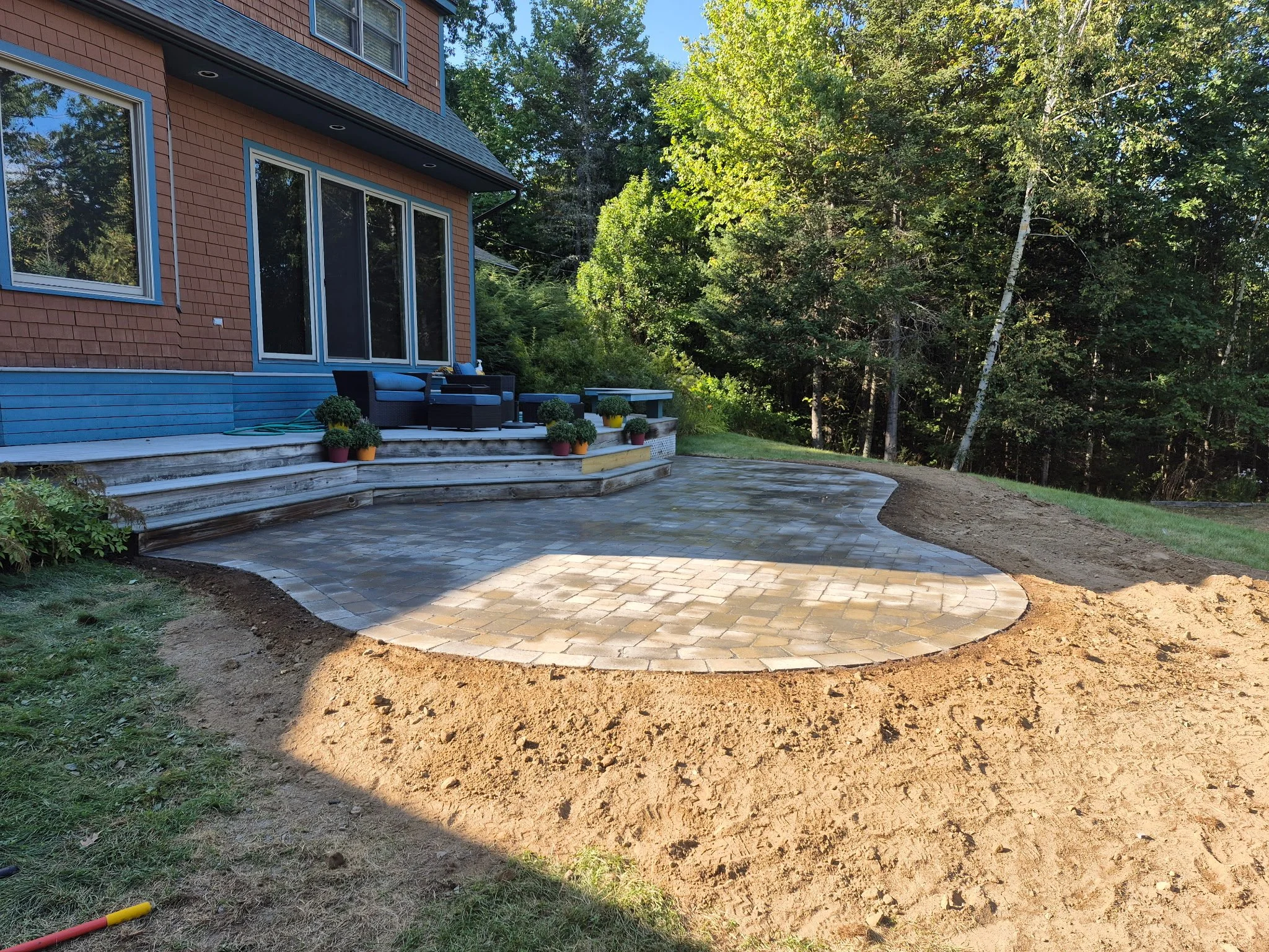 Backyard patio with newly laid pavers, outdoor furniture, potted plants, and a house with large windows and an upper deck, surrounded by trees.