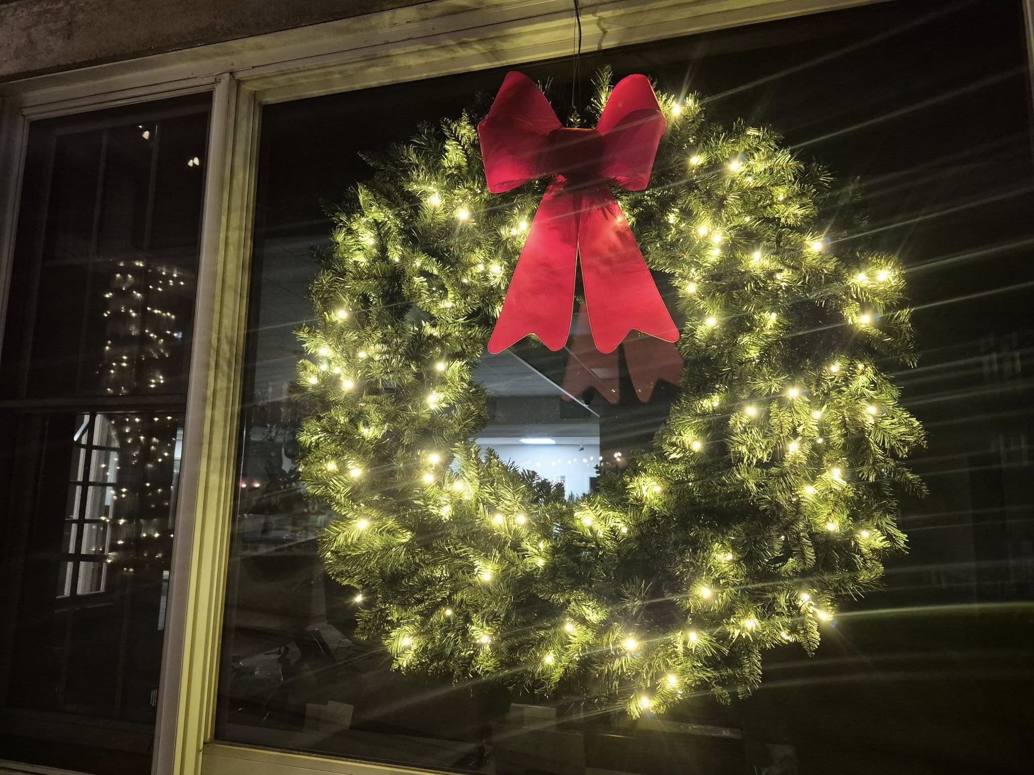 Christmas wreath with lights and a red bow in a window at night.