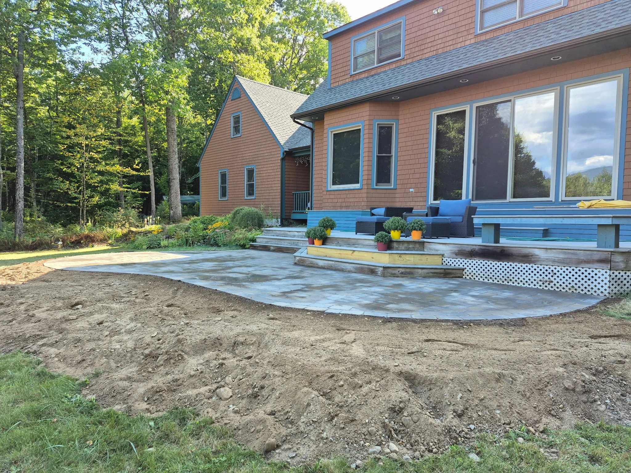 A newly paved patio area with stone pavers, outdoor furniture, and potted plants, adjacent to a house with large glass sliding doors, in a backyard with trees.