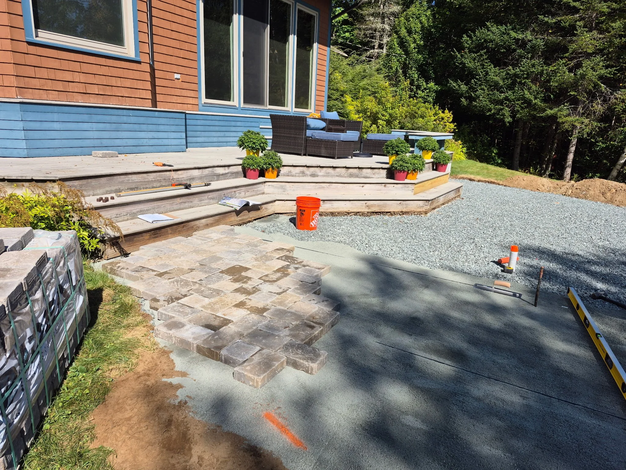 A backyard patio under construction with stone pavers and wooden stairs, outdoor furniture, potted plants, construction tools, and gravel area, surrounded by trees.