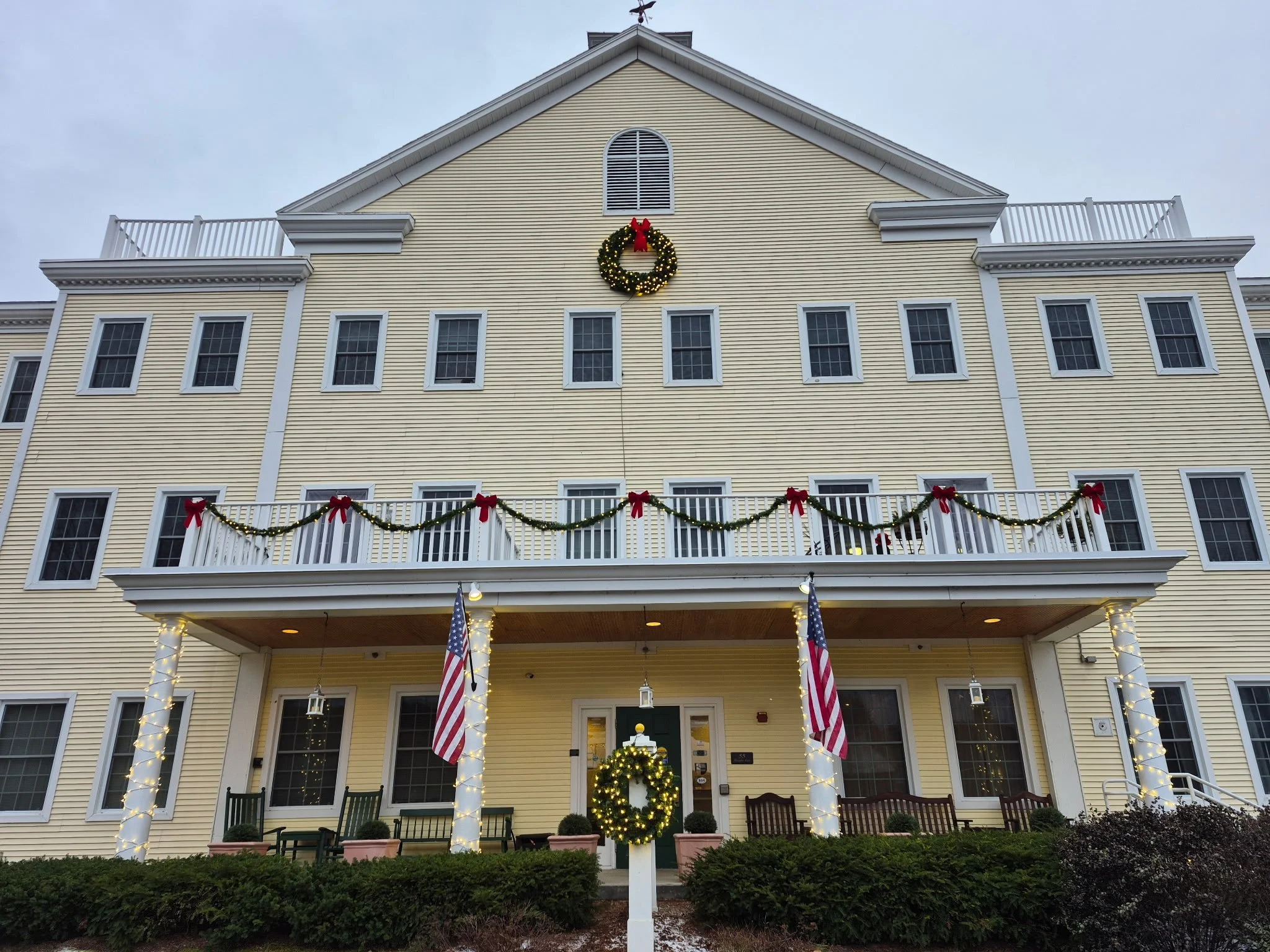 A yellow multi-story building decorated for Christmas with wreaths, garlands, red bows, and American flags. The building has multiple windows, and the porch area is lit with string lights and has benches and plants.