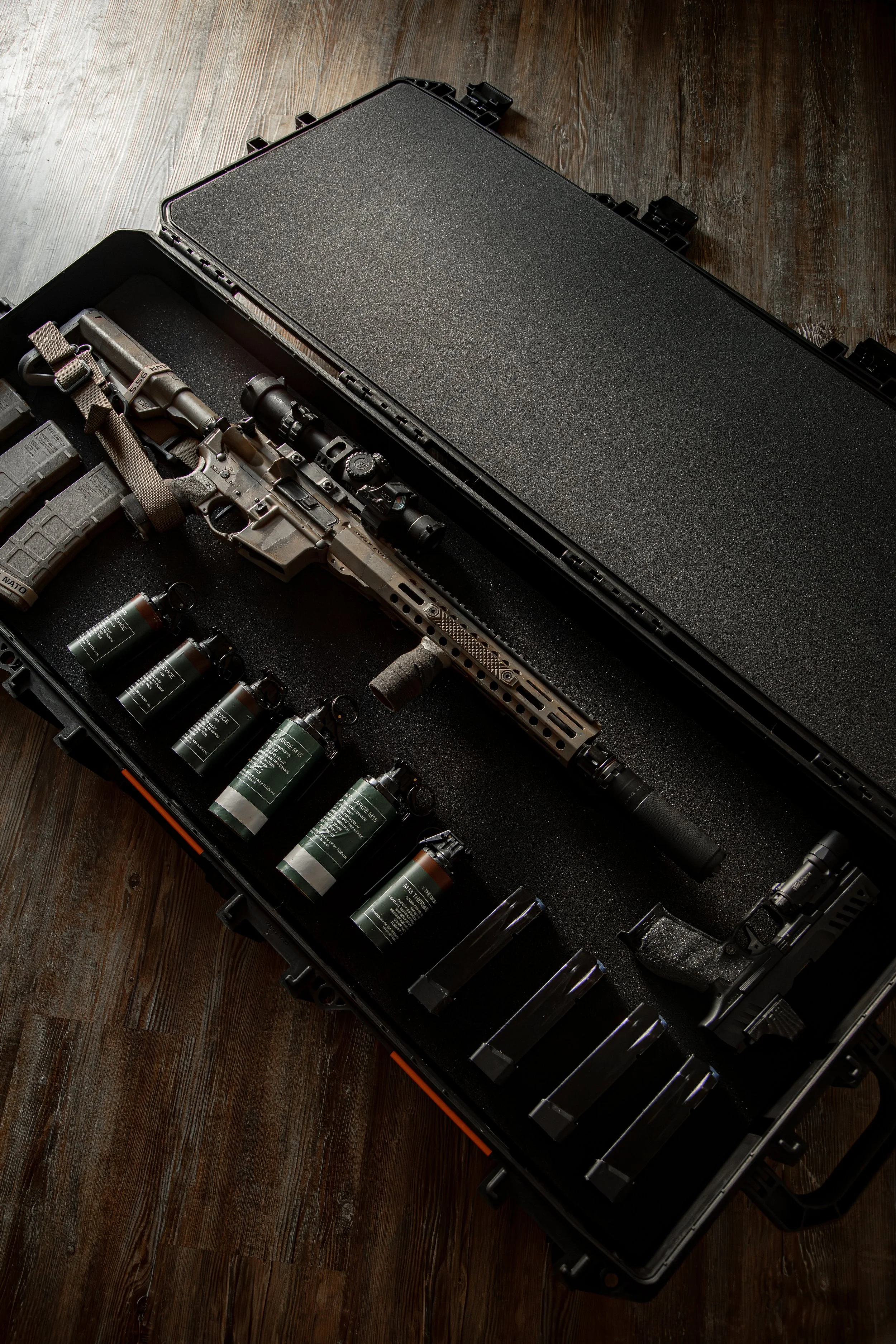 A tactical firearm with a scope, flashlight, and magazines in a black hard case on a wooden floor.