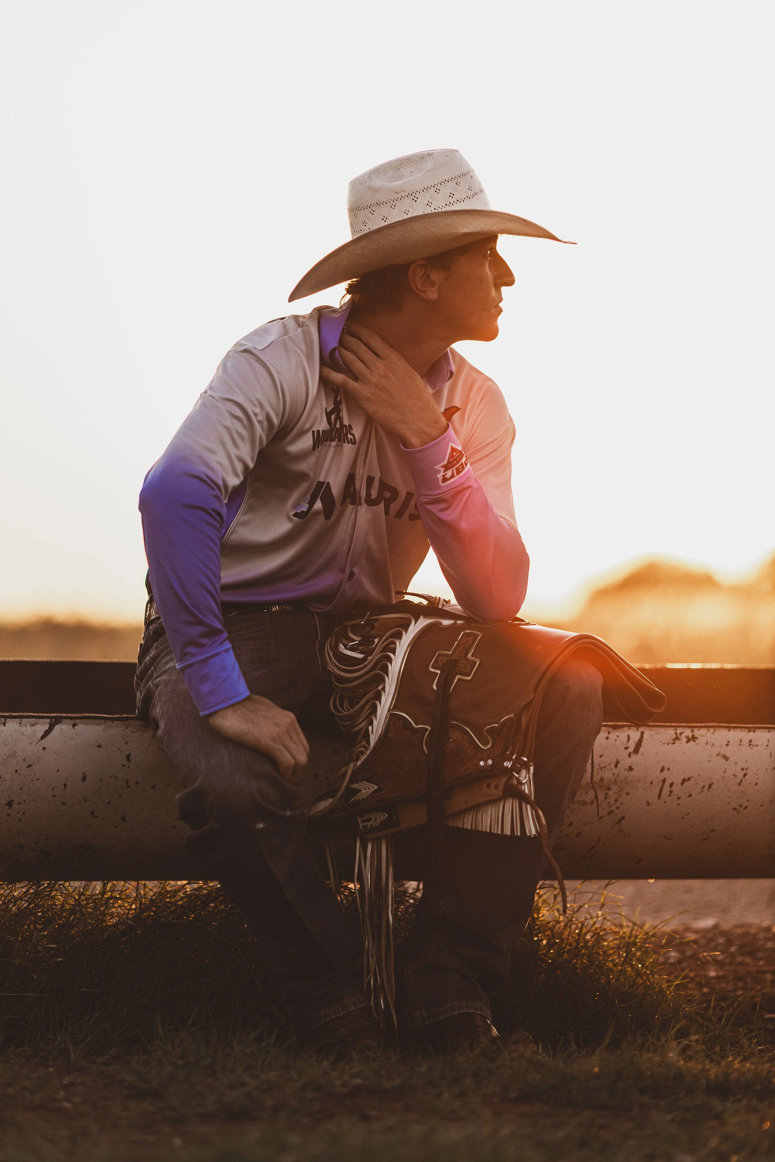 A man wearing a cowboy hat, pink and purple shirt, and jeans sitting on a fence at sunset with a baseball glove and baseball bat.