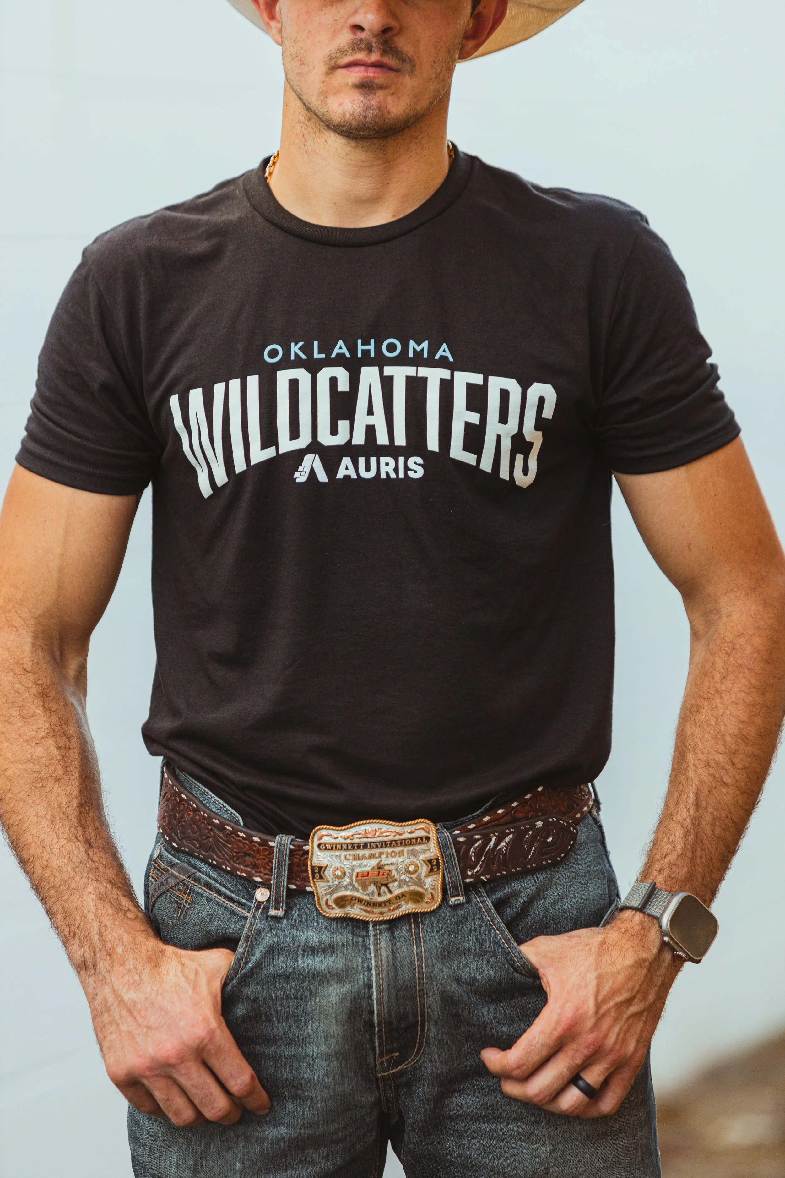 Man wearing a cowboy hat, black 'Oklahoma Wildcatters' t-shirt, jeans, and large belt buckle, standing with crossed hands.