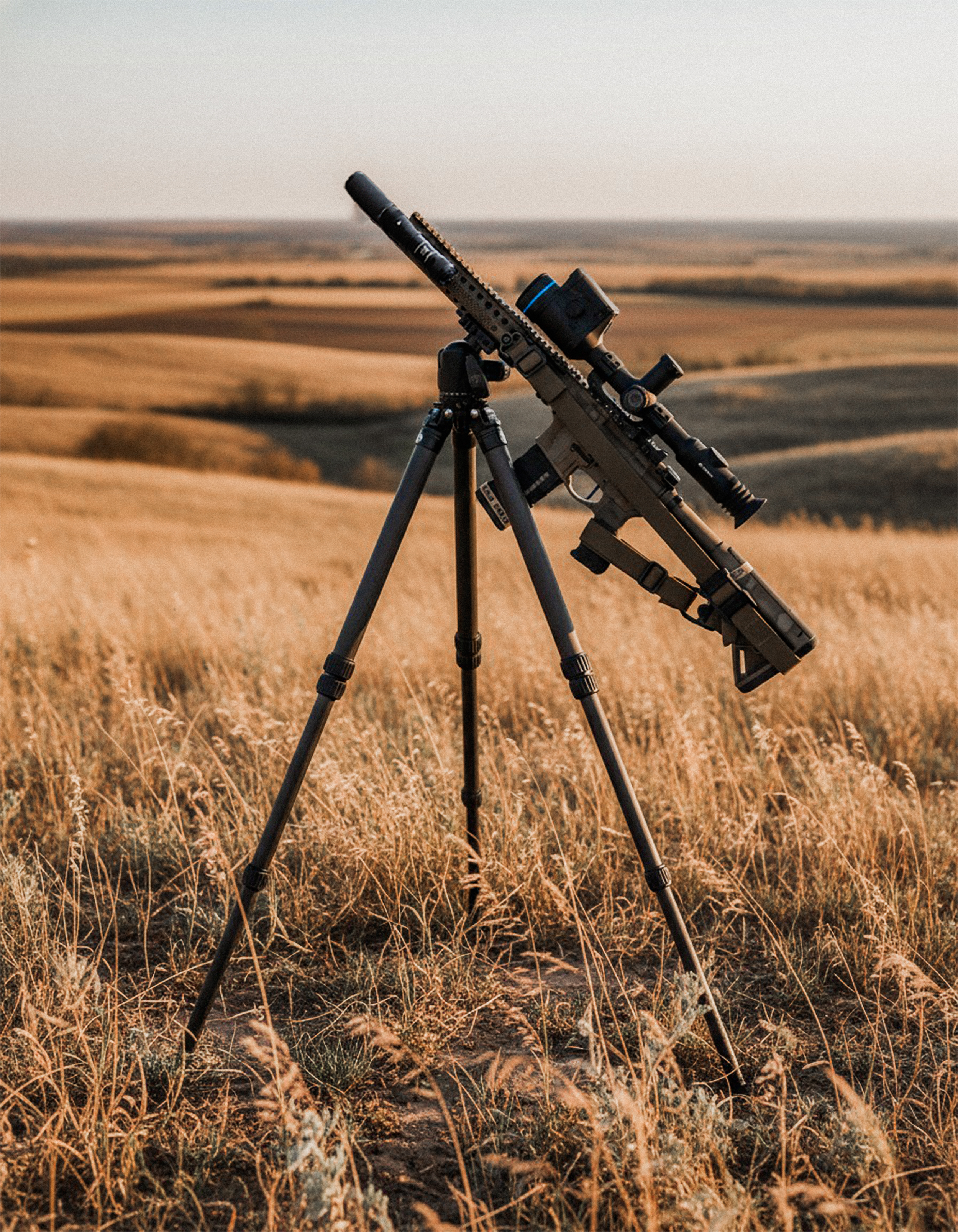A sniper rifle with a scope mounted on a tripod in a grassy field during sunset or sunrise.