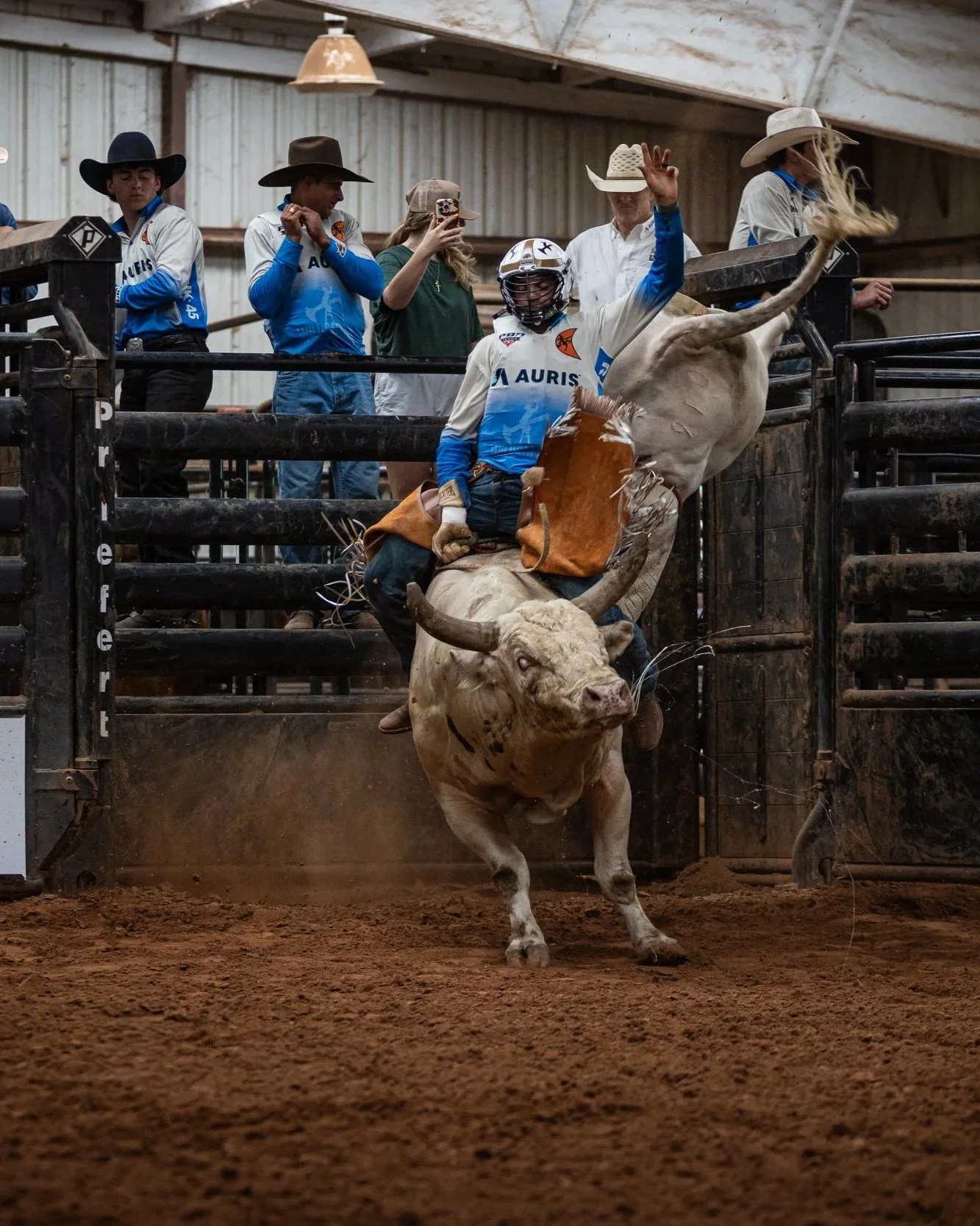 A rodeo event where a cowboy is riding a bucking bull inside an arena, with spectators and other rodeo participants watching from the sidelines.