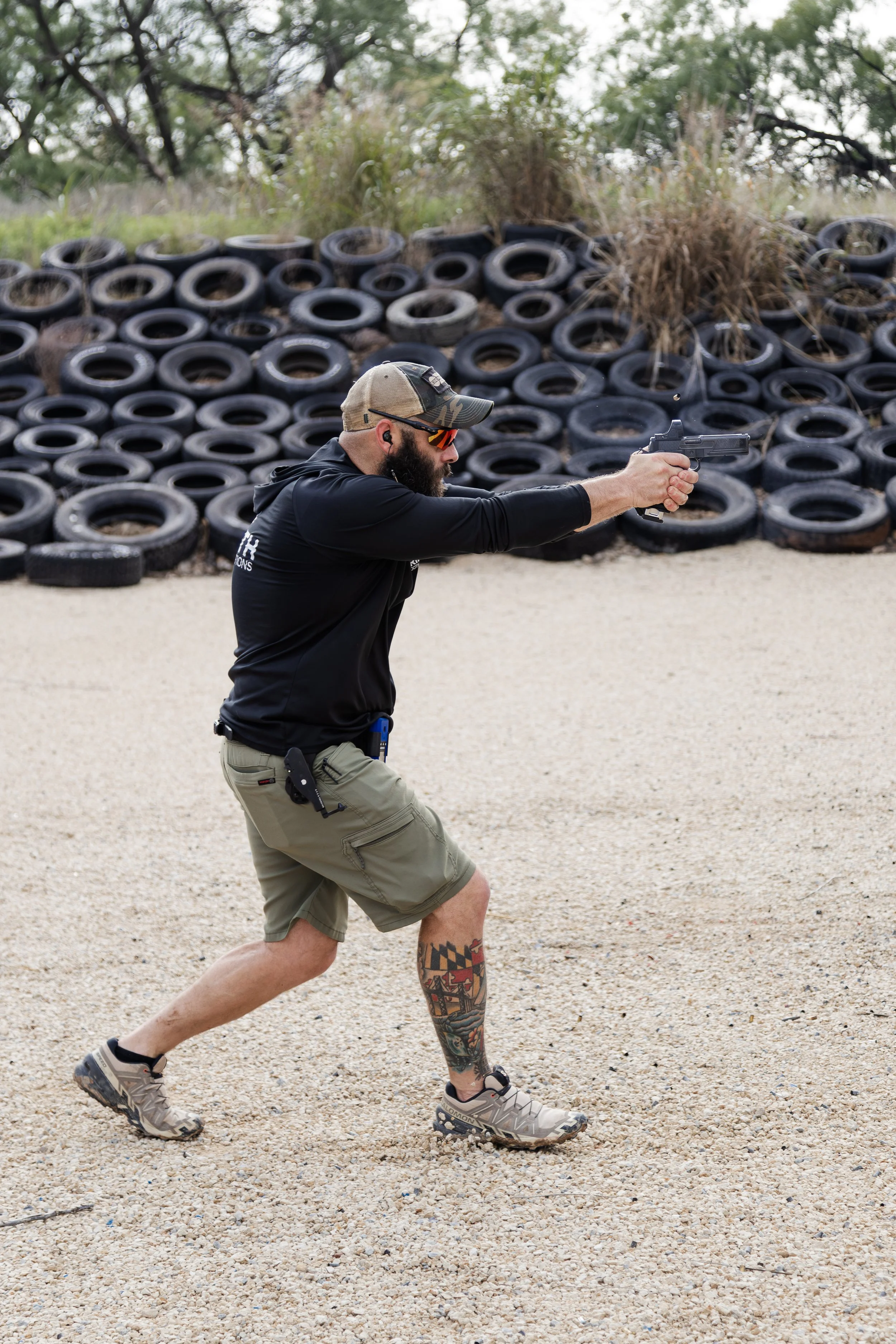 A man with a beard, tattooed leg, wearing a tan cap, sunglasses, black shirt, khaki shorts, and sneakers is engaged in shooting with a handgun at a shooting range. He holds the gun with both hands and is aiming forward. Behind him, there is a large pile of old tires and trees.