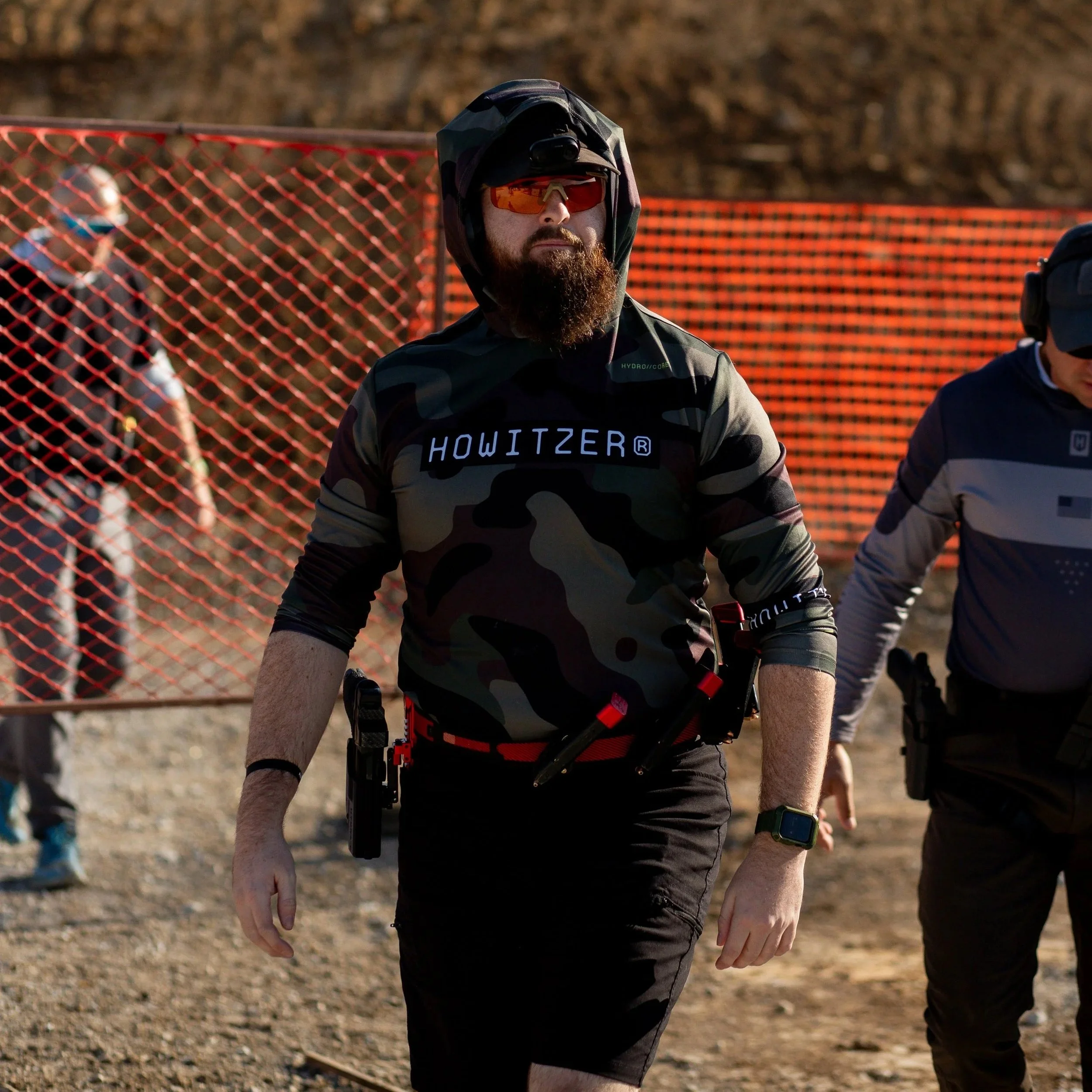 A man with a beard wearing sunglasses, a camouflage hoodie with 'HOWITZER' written on it, and black shorts, walking outdoors with others in a shooting range or similar setting.