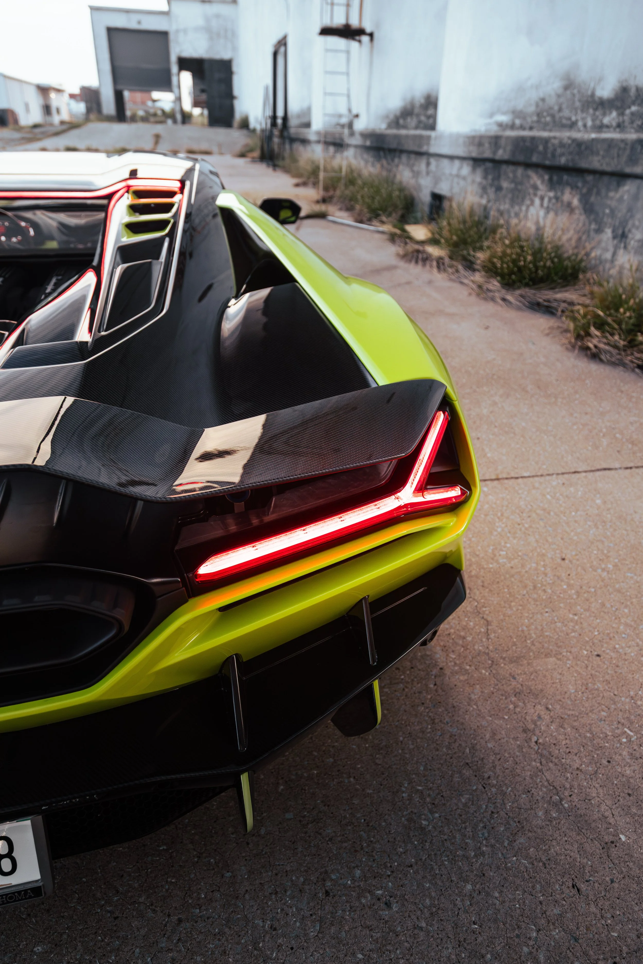 Close-up of the rear end of a bright green sports car with carbon fiber details, parked on a sandy, paved area in front of a weathered, white building.