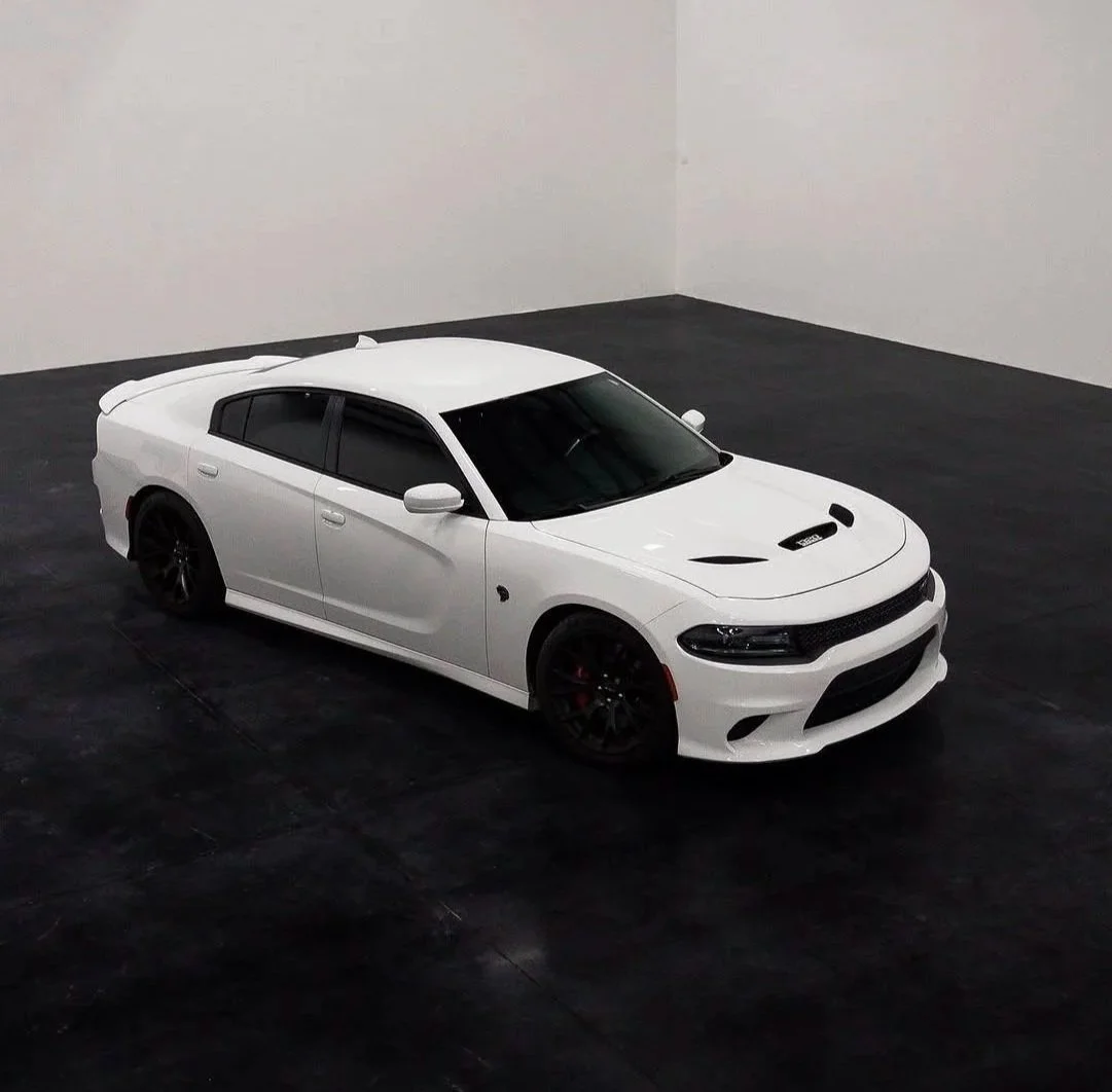 A white sports sedan with black wheels parked in a plain indoor showroom with black flooring and white walls.