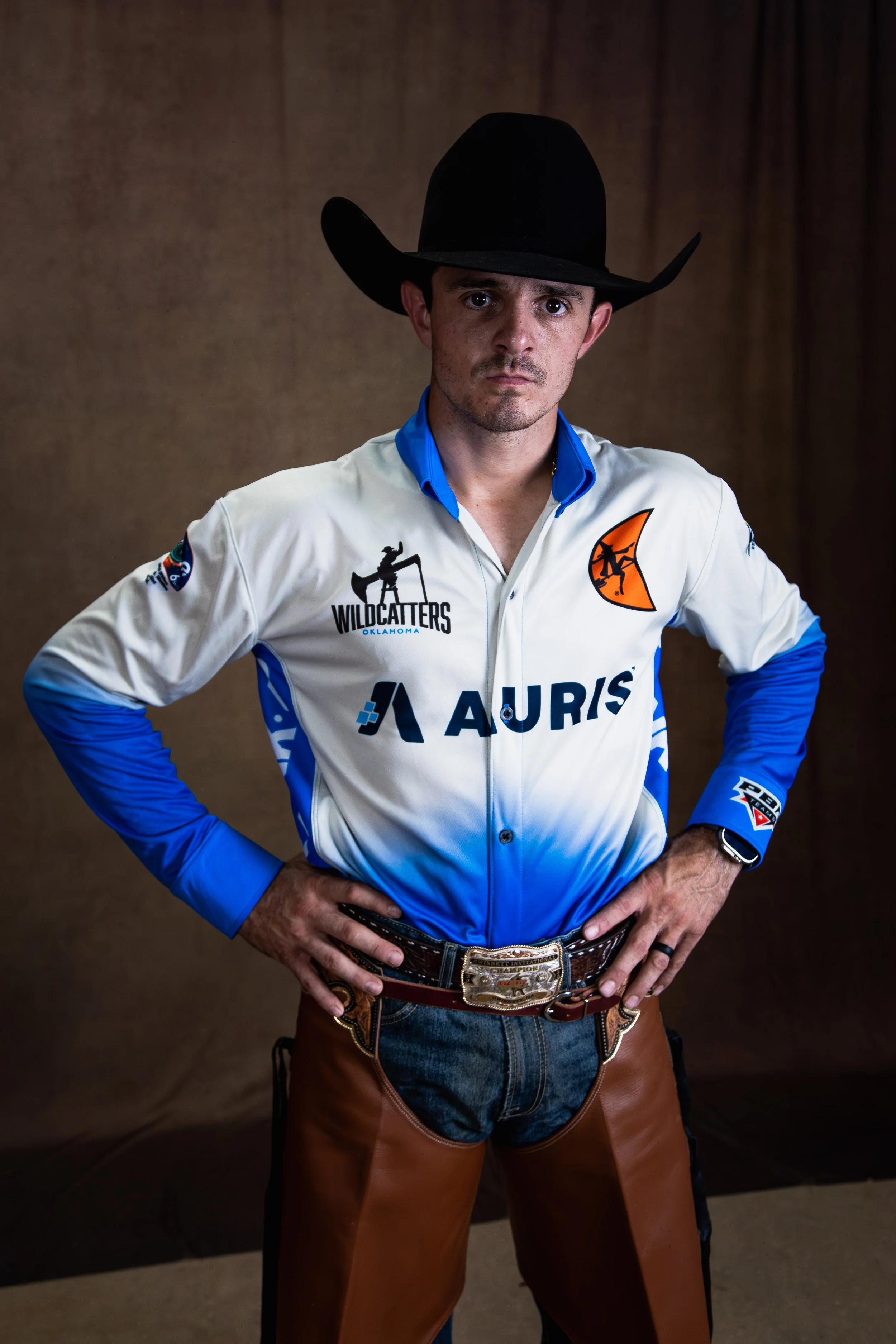 A man wearing a black cowboy hat, a white and blue ranching jacket, a large belt buckle, and brown leather chaps, standing with hands on his hips against a plain background.
