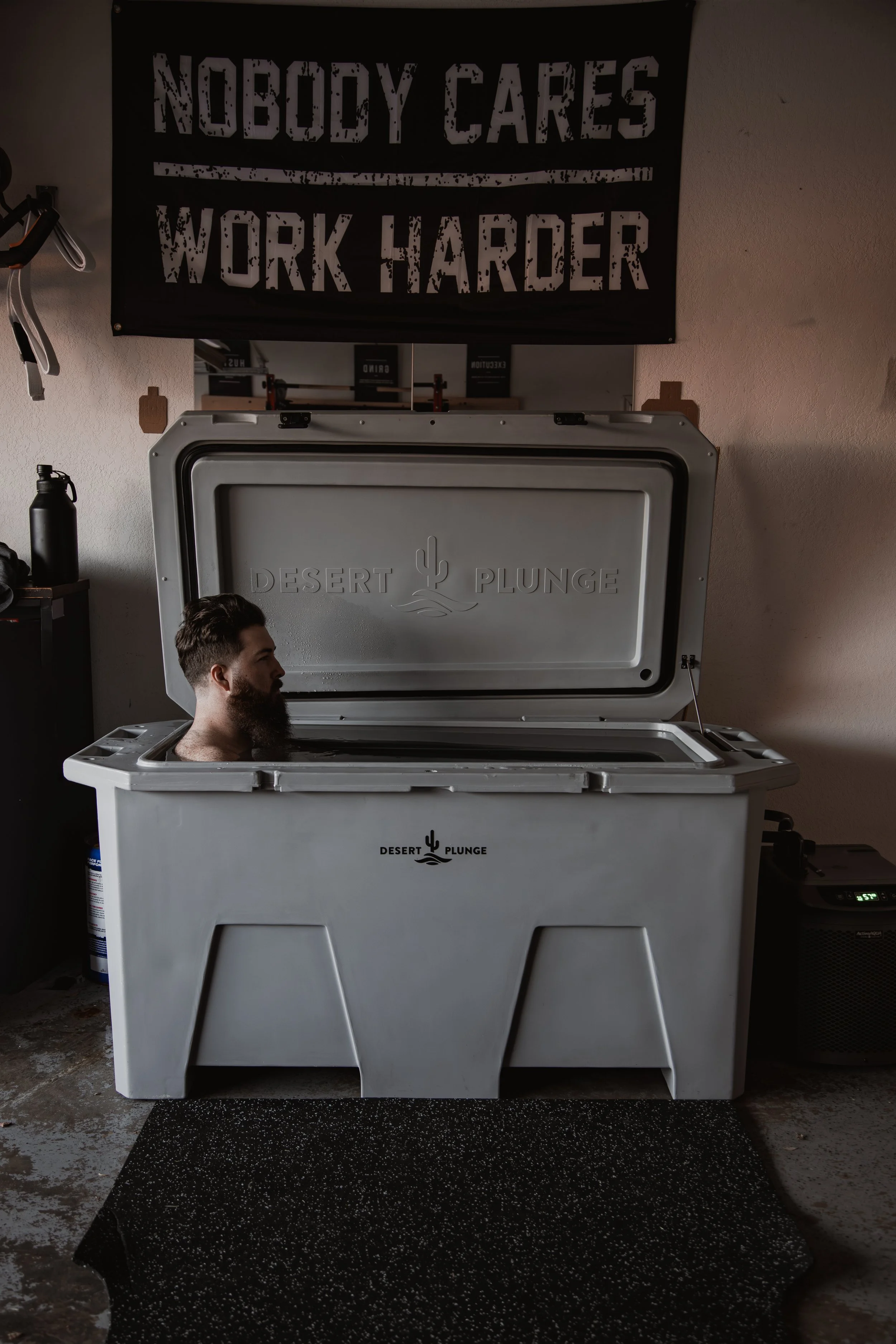 A man with a beard relaxing in a Desert Plunge immersion tub in a fitness or wellness space, with a motivational sign reading 'Nobody Cares Work Harder' hanging on the wall above.