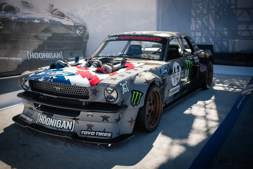 A race car with an American flag-themed paint job, sponsor logos including Ford and Monster Energy, displayed at an auto show with shadows cast on the floor.