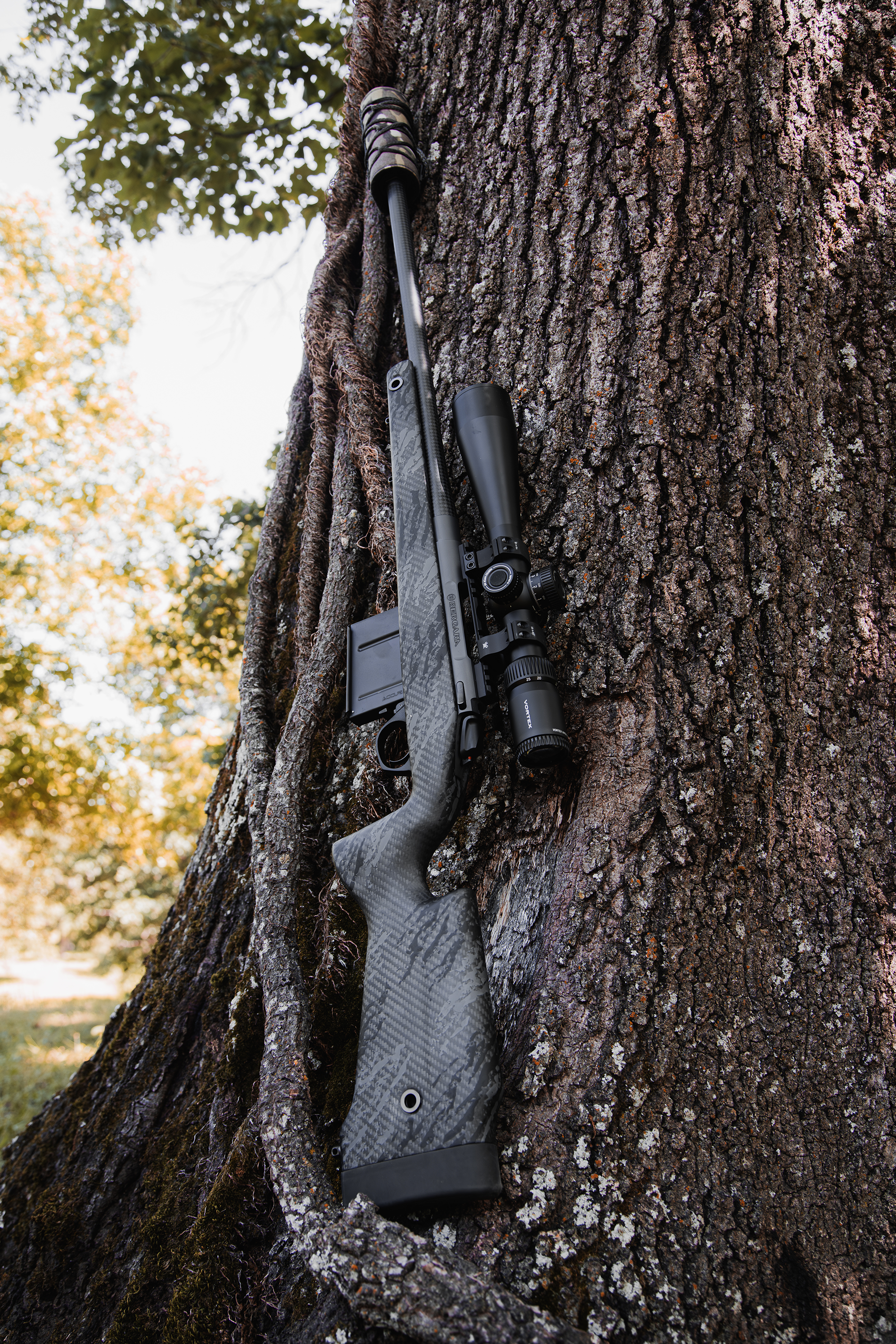 A hunting rifle with a scope mounted on it is leaning against a large tree trunk in an outdoor setting, with blurred trees and sunlight in the background.