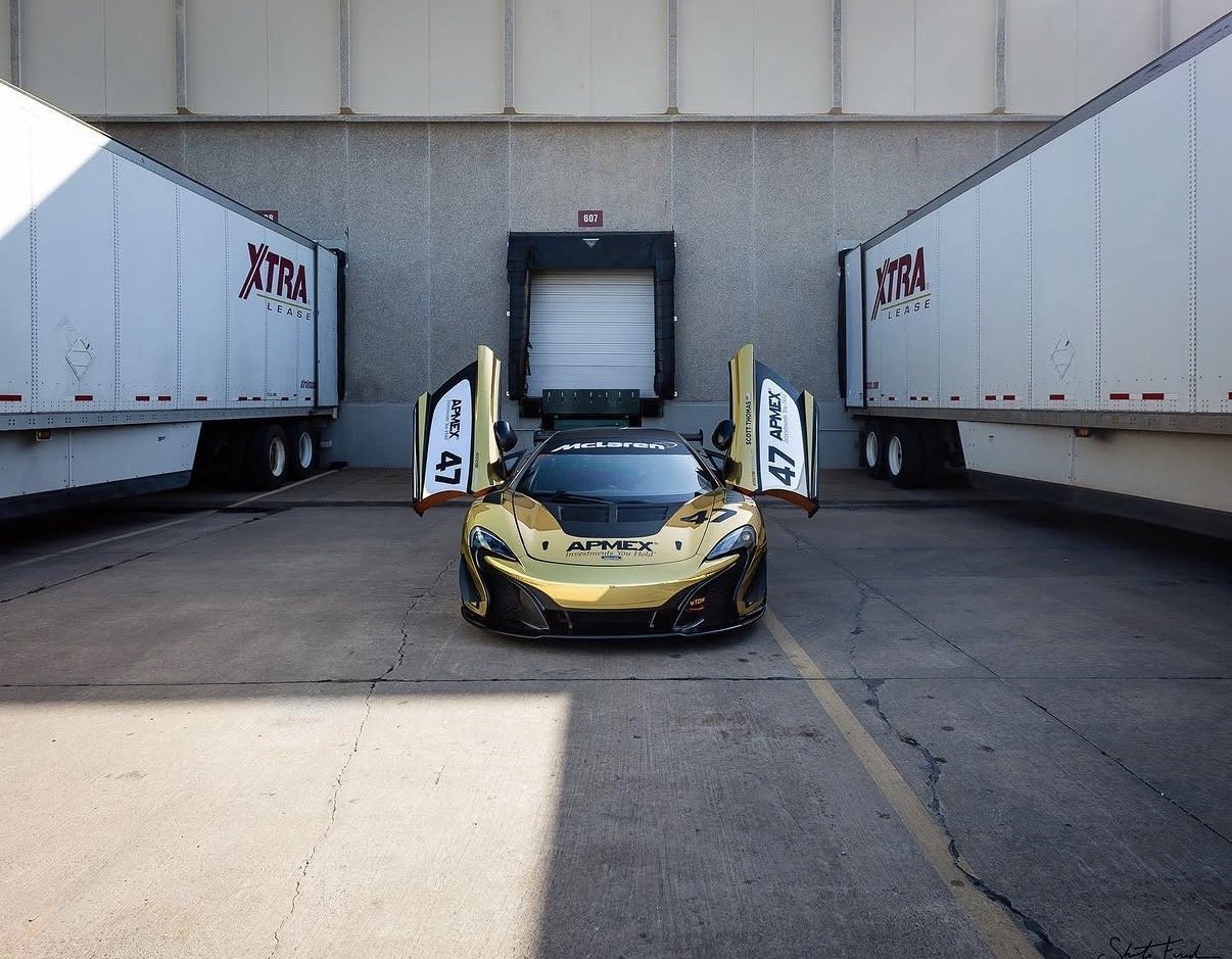 A gold and black McLaren race car with its doors open, parked between two large semi-trailers marked 'XTRA Lease' at a loading dock.