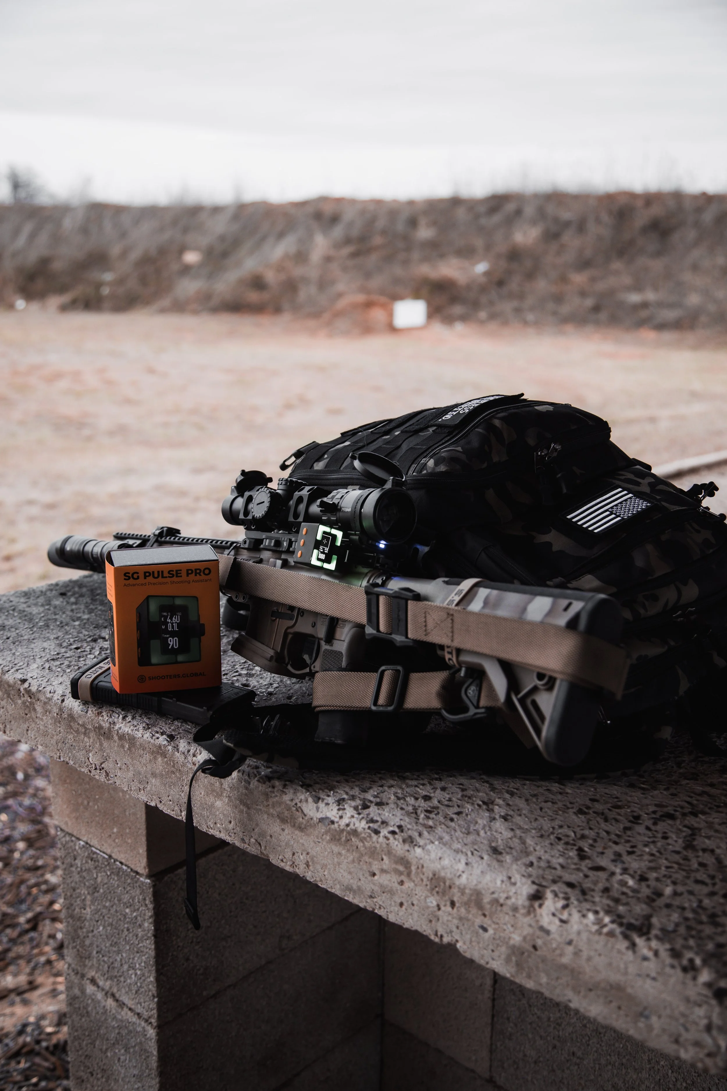 A rifle with a scope mounted on it resting on a concrete block with a handheld device and orange box labeled SG Pulse Pro nearby. Background is a barren outdoor landscape with dirt and a cliff.