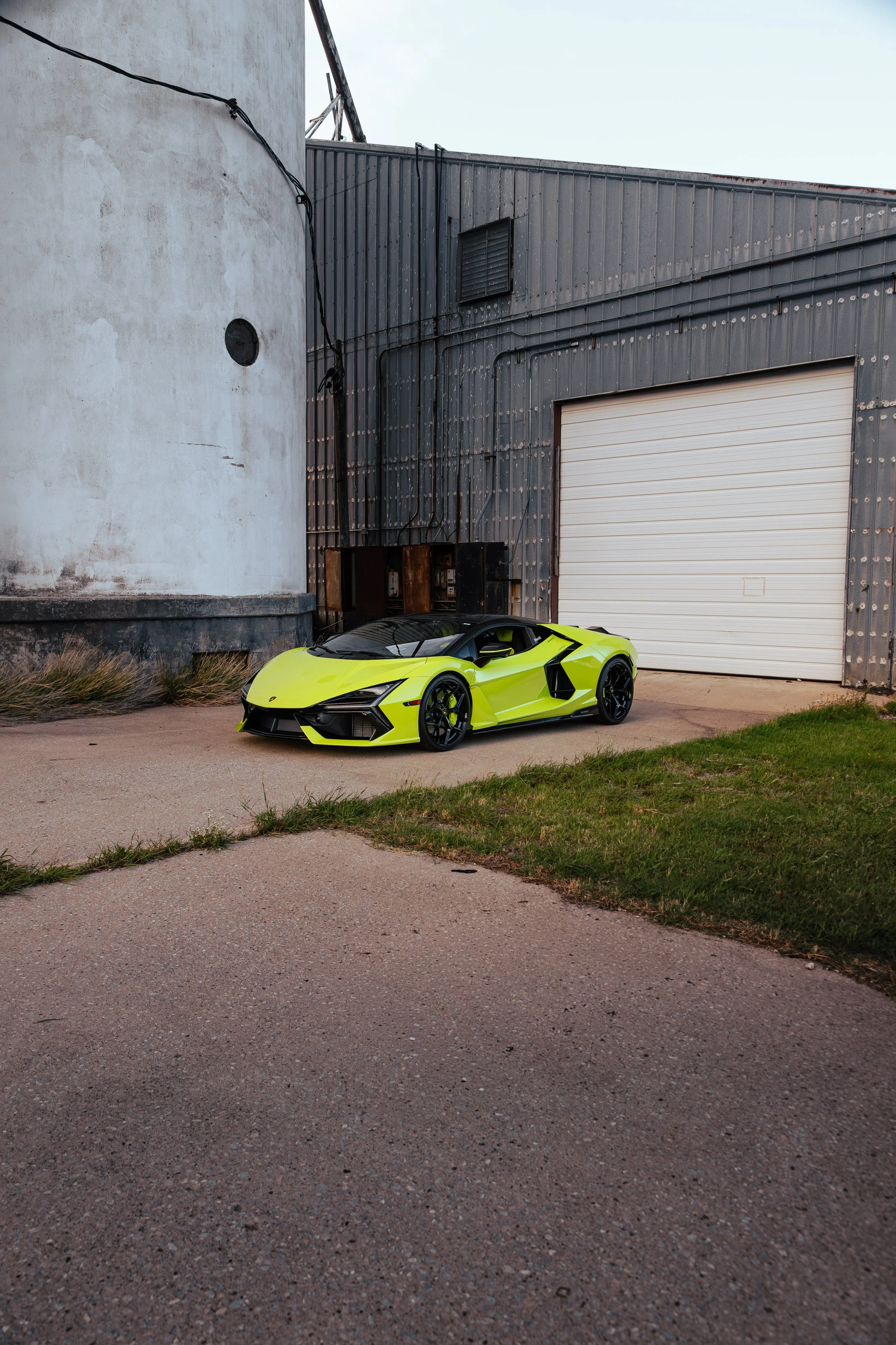 A bright yellow sports car parked against a textured concrete and metal industrial building with a large white garage door, some green grass, and a small grassy area in the foreground.
