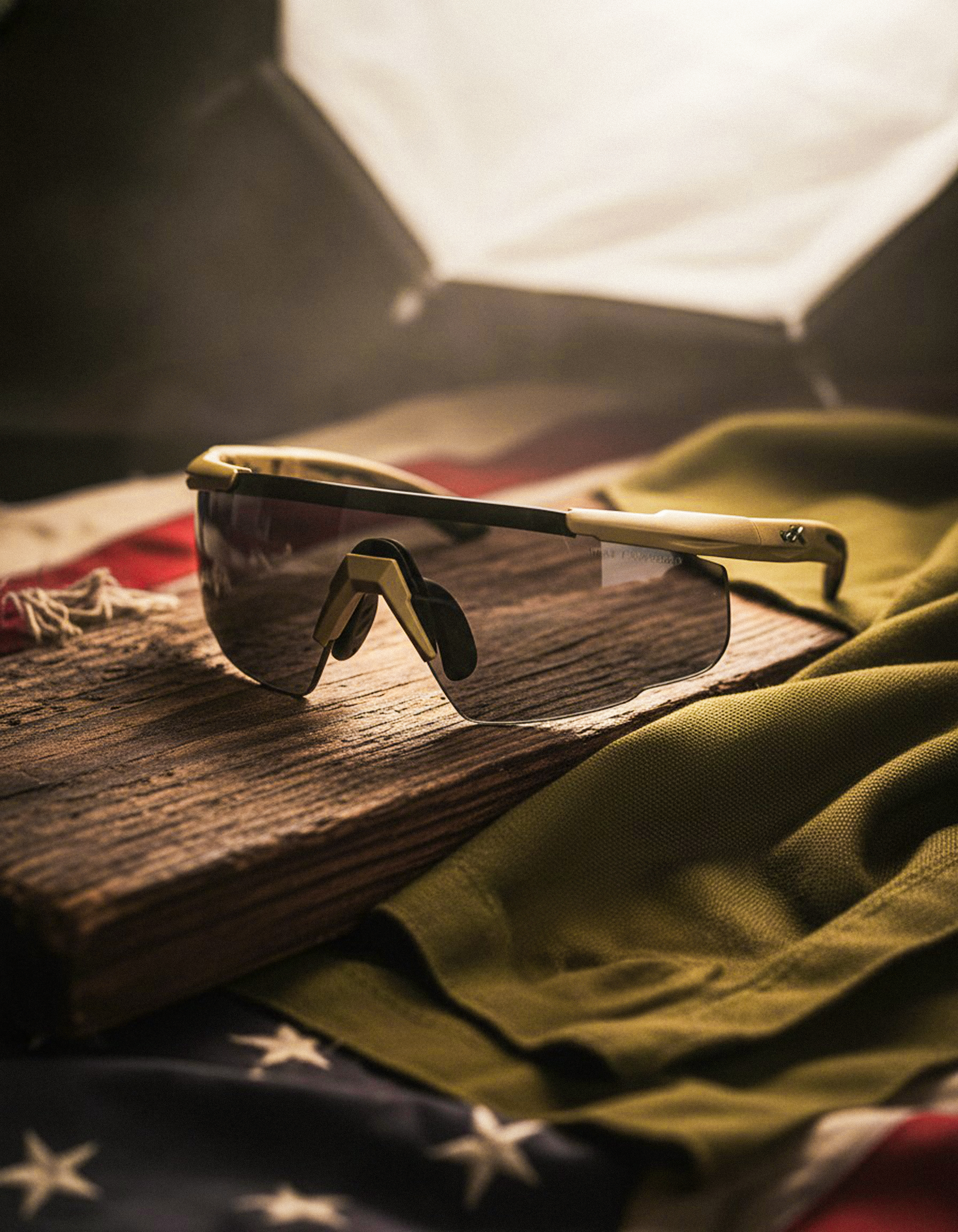 A pair of sports sunglasses with dark lenses resting on a wooden surface surrounded by fabric pieces, including a green cloth and an American flag with stars.