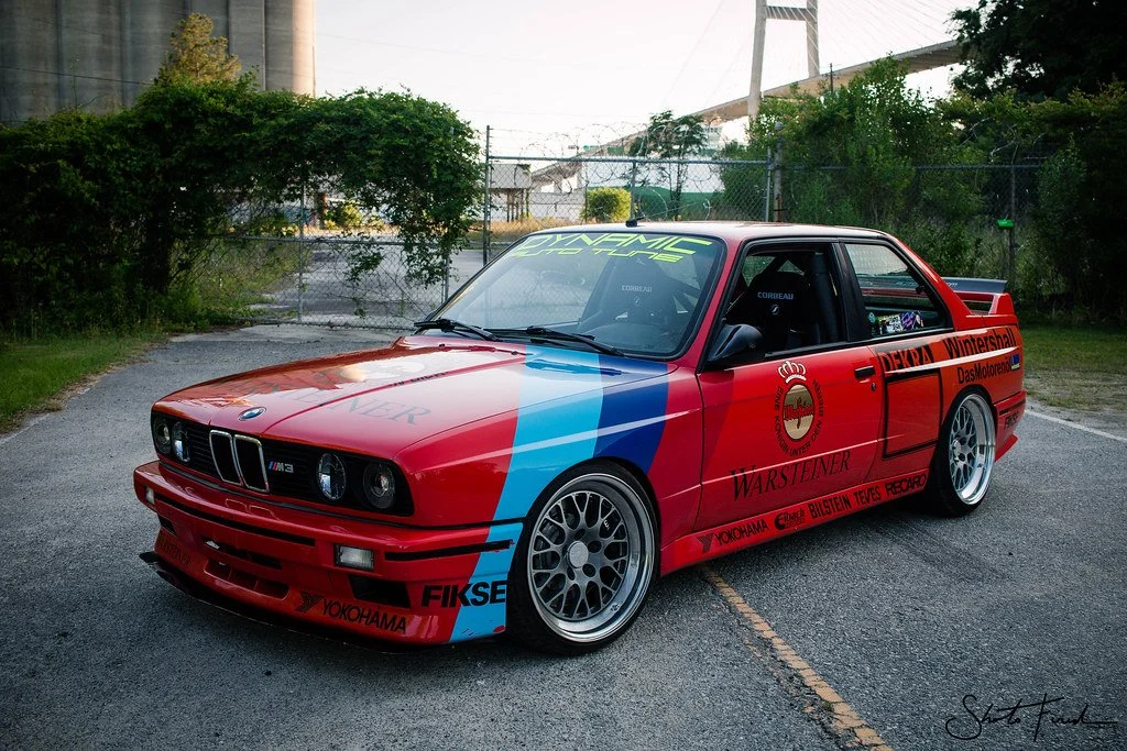 Red and blue race car with sponsor decals and aftermarket wheels parked on a paved lot with a chain-link fence and industrial background.