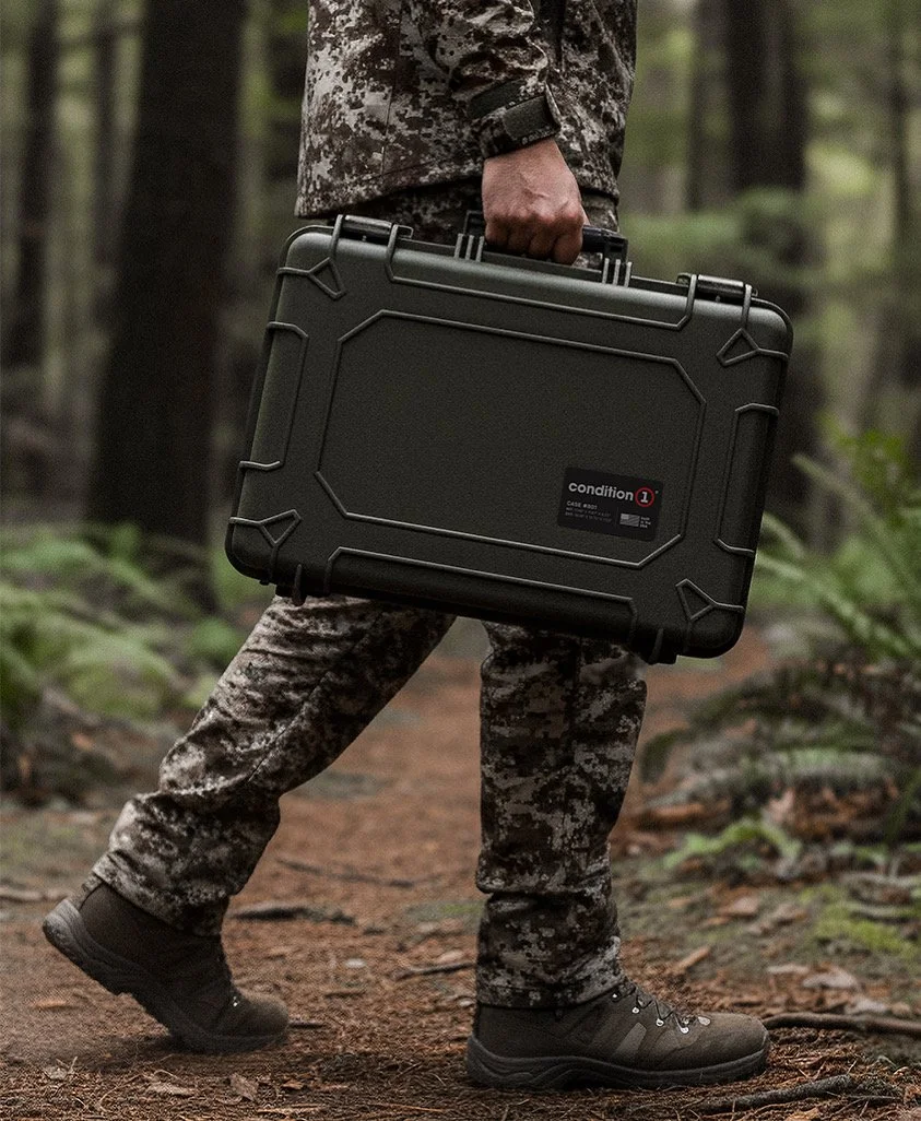 Person in camouflage clothing walking through a forest, carrying a black hard case in their right hand.