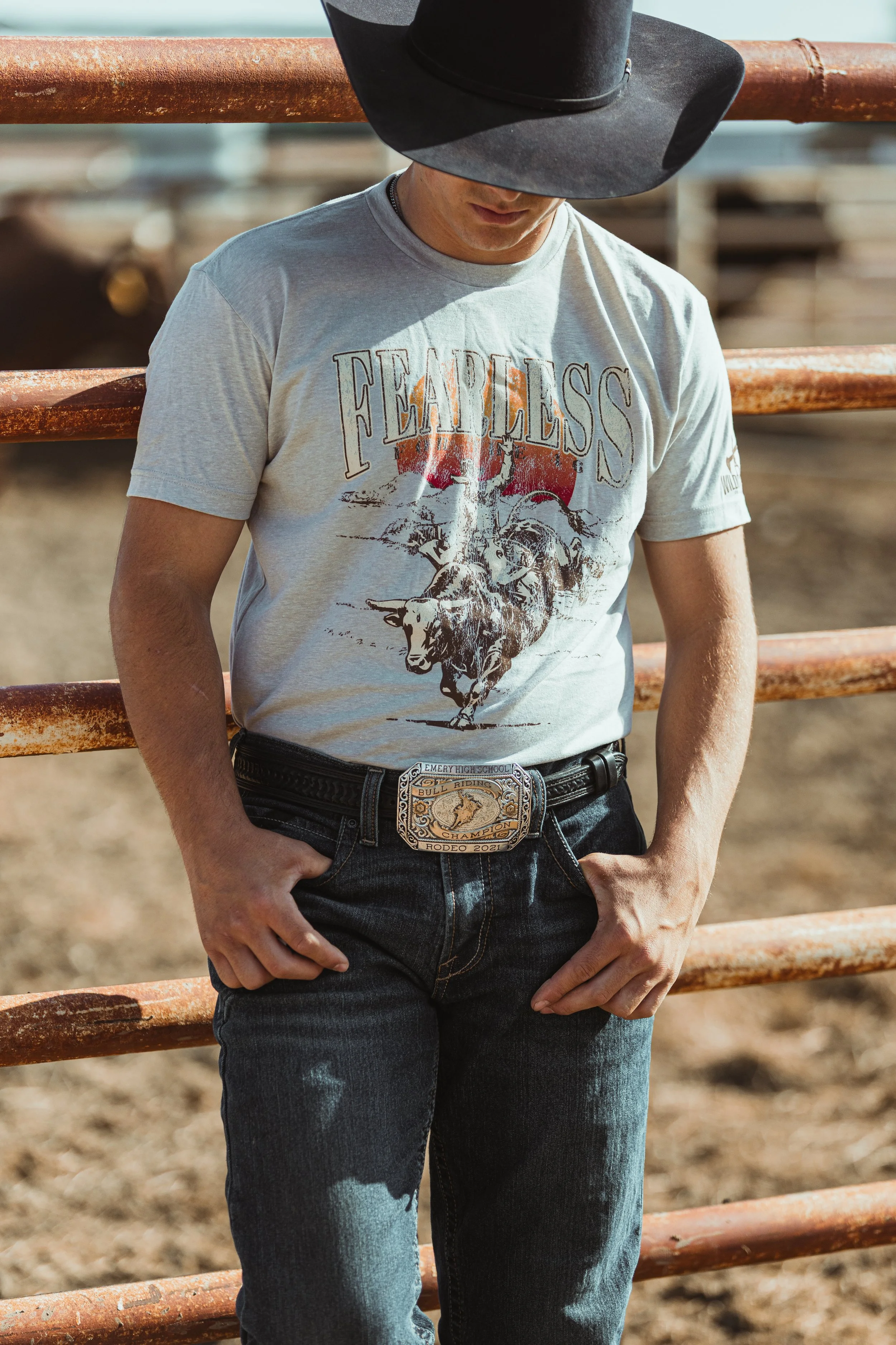A man wearing a black cowboy hat, a gray graphic T-shirt with a bull riding scene, and jeans with a large belt buckle, standing outdoors next to rusted metal fencing.