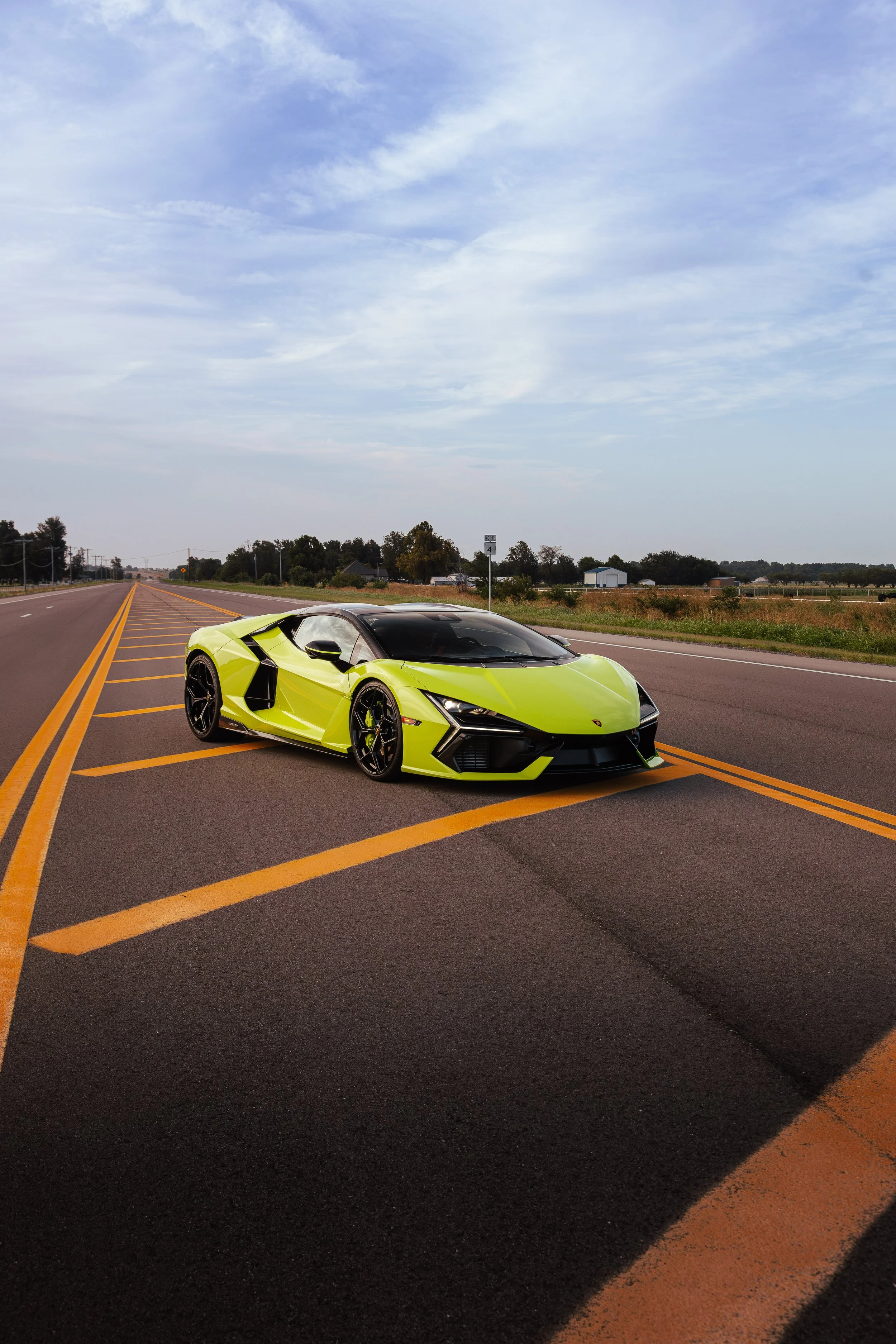 A bright yellow sports car parked on the side of a two-lane road under a partly cloudy sky, with fields and farm buildings in the background.