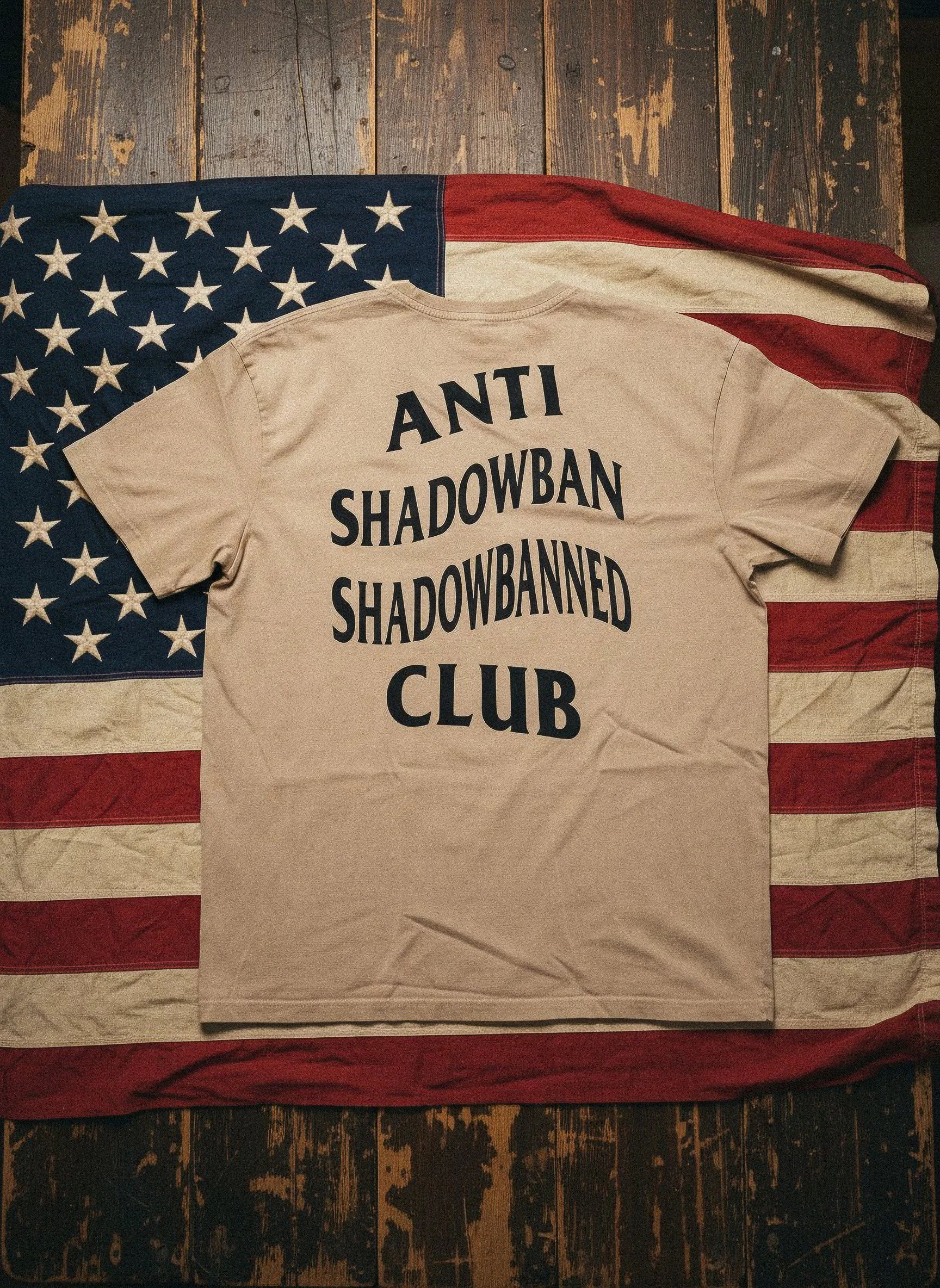 A beige T-shirt with black text that says 'Anti Shadowban Club,' lying on top of an American flag draped over a wooden surface.