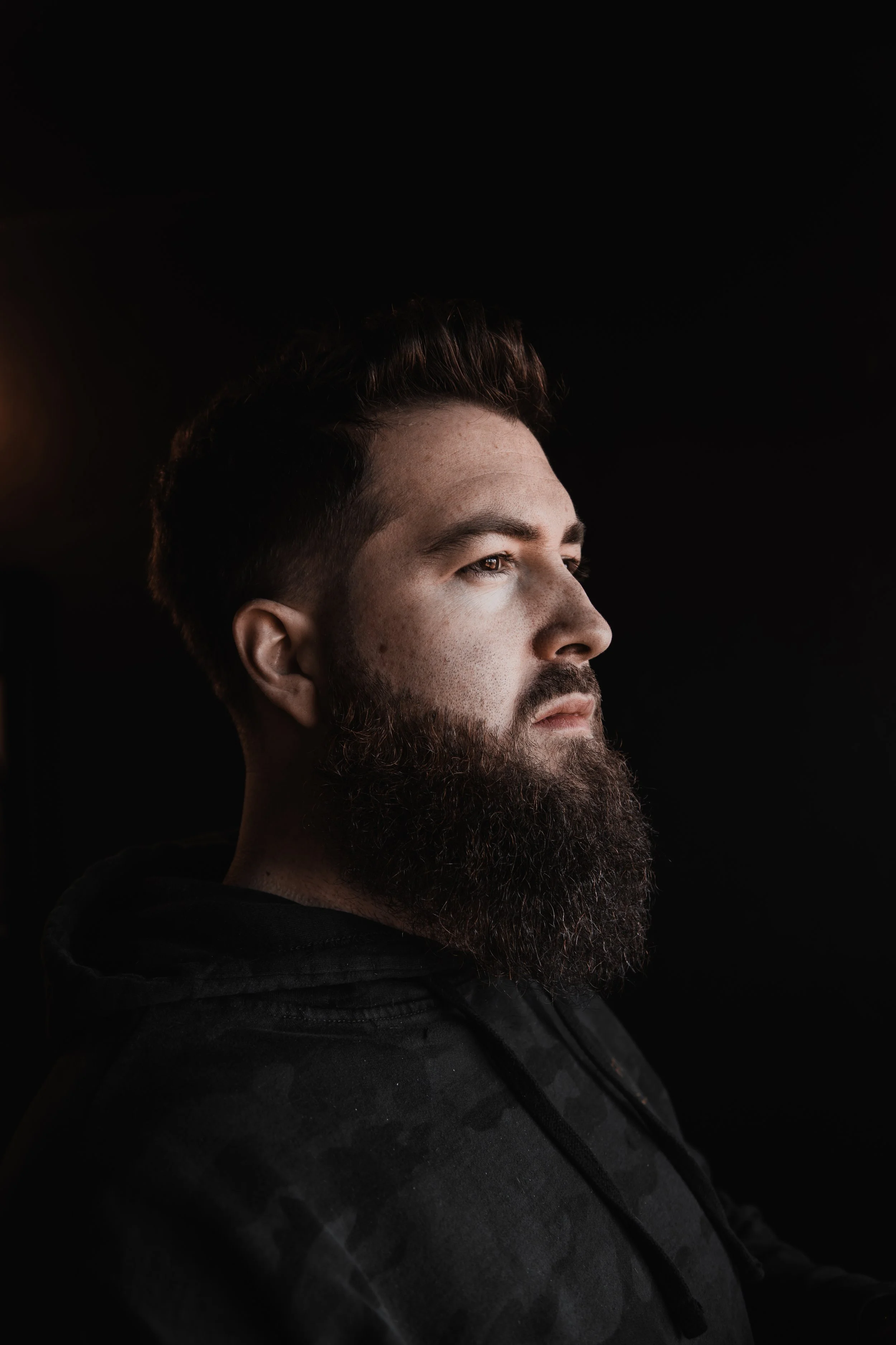 Side profile of a bearded man with short hair wearing a dark hoodie against a black background.