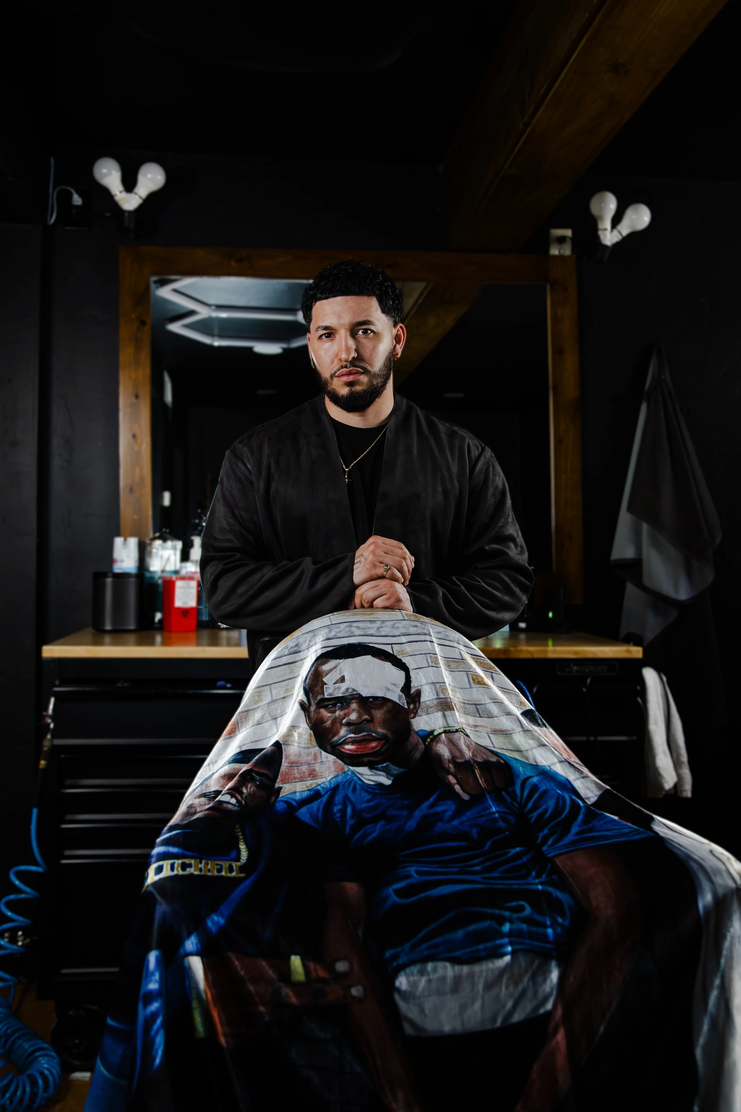 A man with dark hair and a beard, wearing a black jacket and gold chain, standing in a barbershop or salon behind a barber chair with a haircut cape depicting a person making a peace sign.