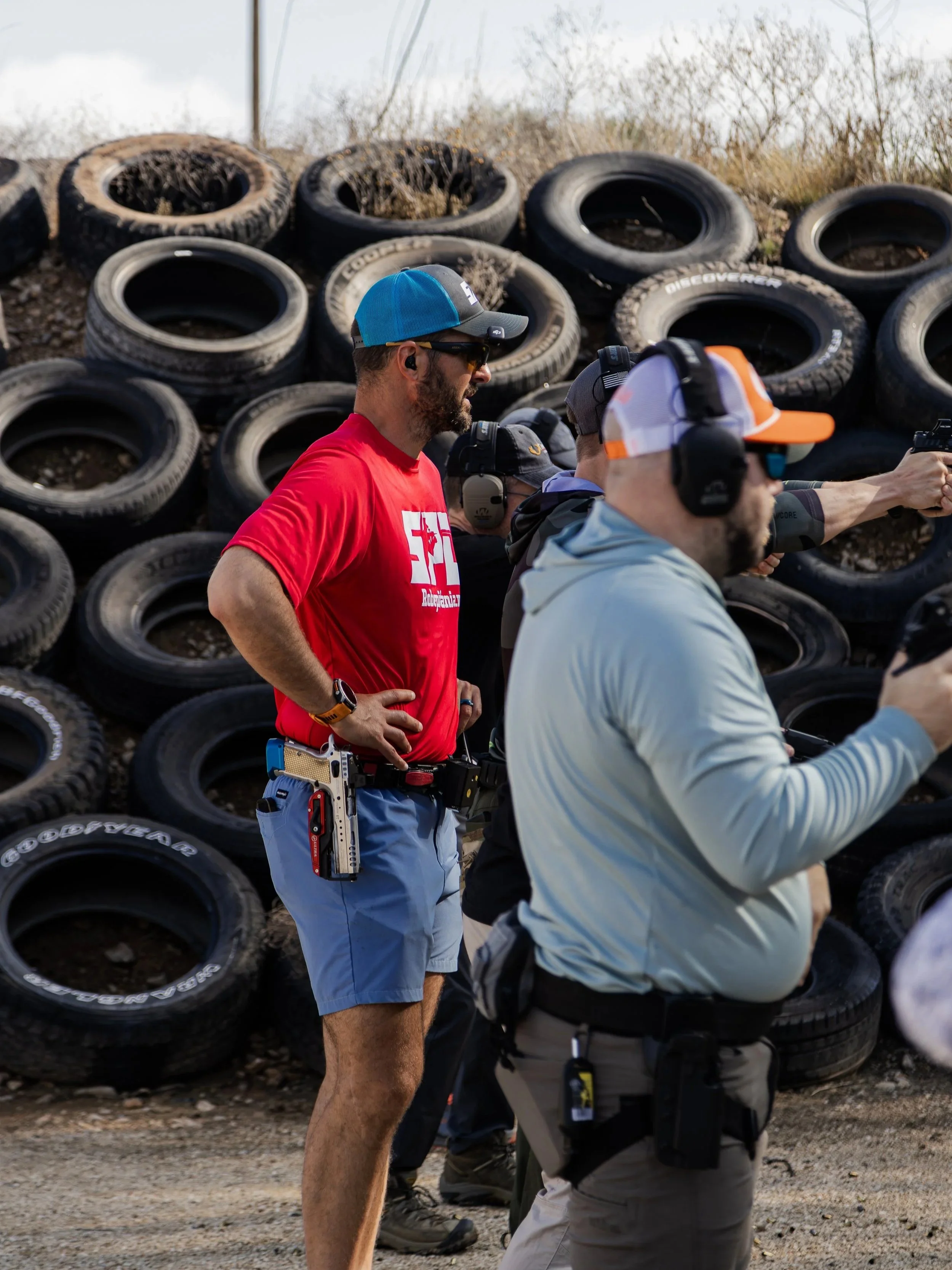 Group of people at a shooting range, with one person aiming a handgun and others standing nearby, against a backdrop of stacked tires.