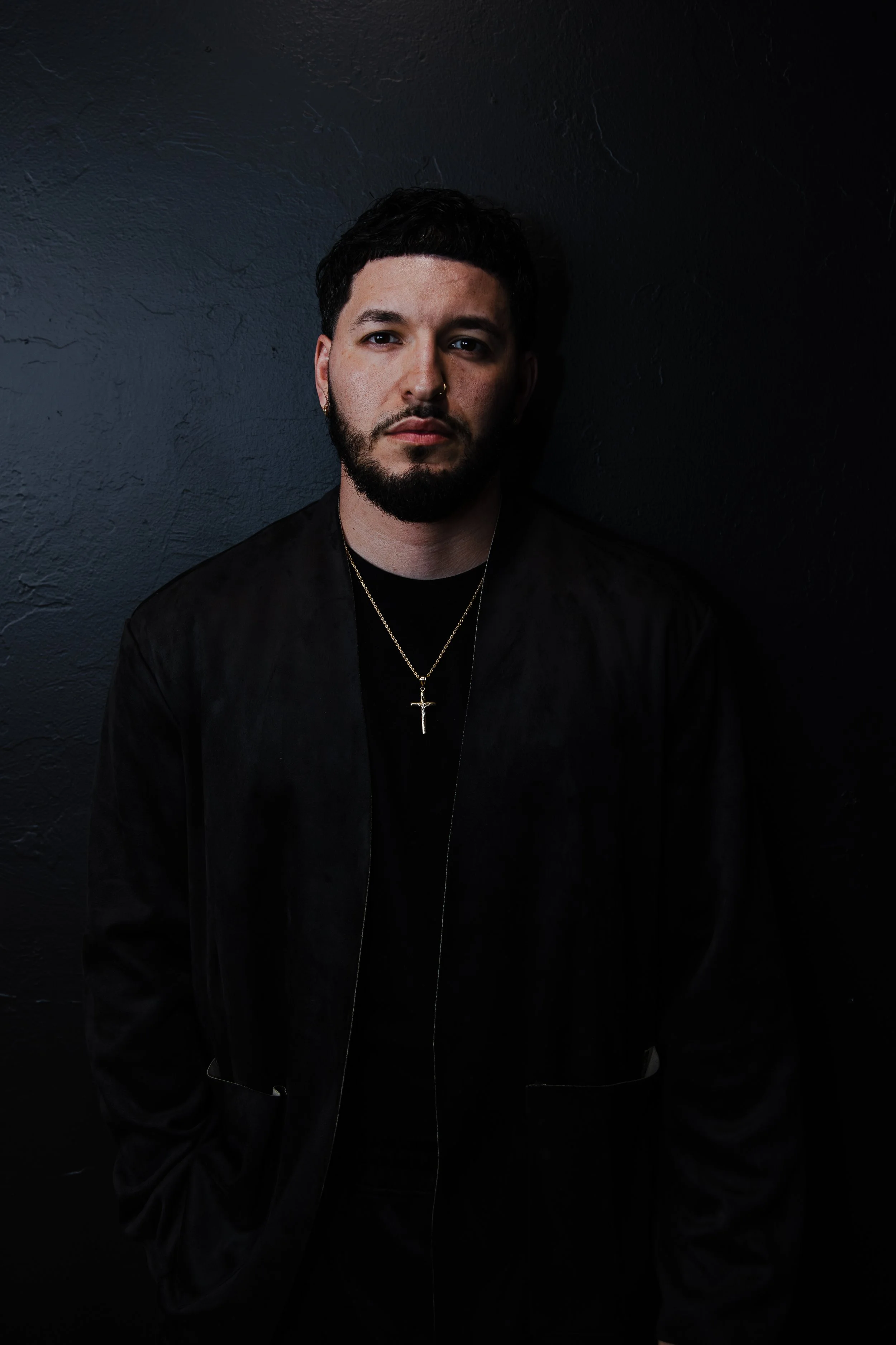 Portrait of a man with dark hair and beard, wearing a black jacket and gold cross necklace, standing against a dark wall.