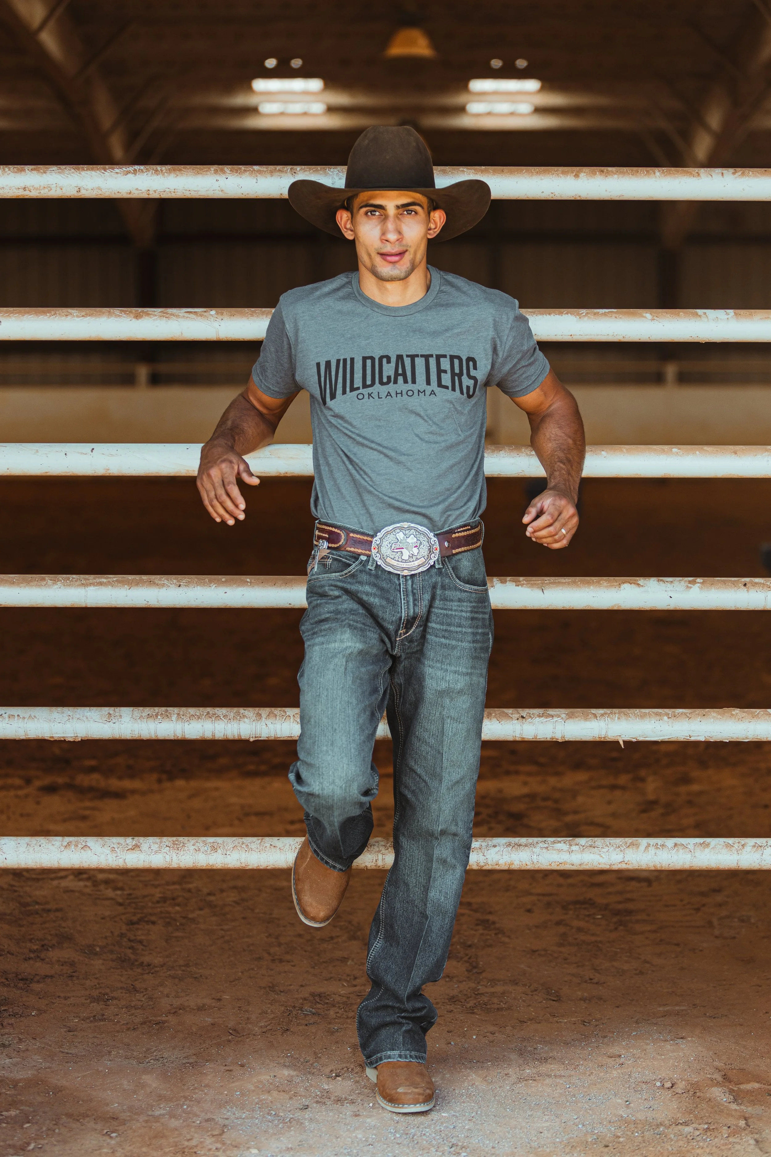 A young man with dark hair wearing a cowboy hat, fitted gray T-shirt with 'WILDCATTERS OKLAHOMA' printed on it, jeans, and brown cowboy boots, leaning against a white metal fence inside a wooden barn or arena with dirt ground.