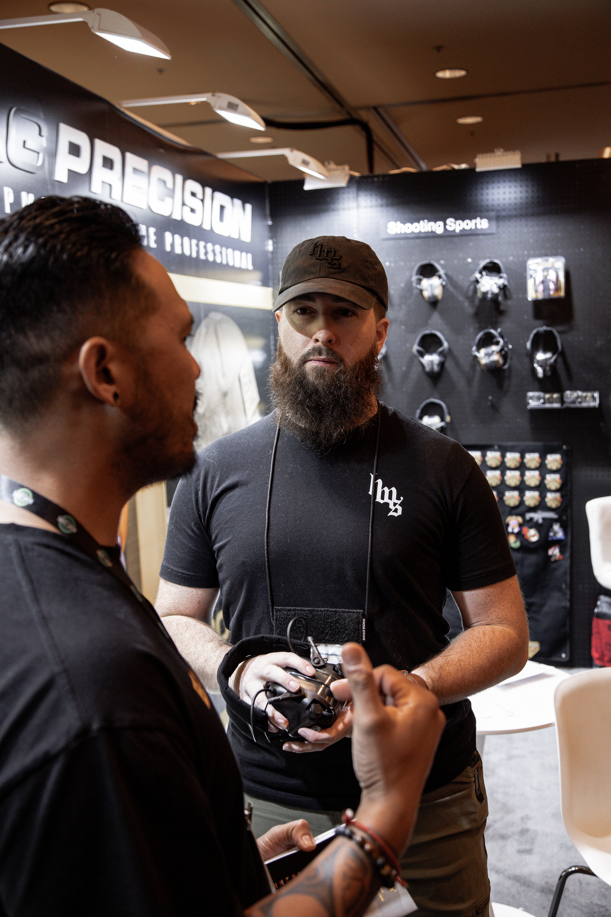 Two men having a conversation at a trade show booth for shooting sports and precision equipment, with one man holding a camera and the other man gesturing.