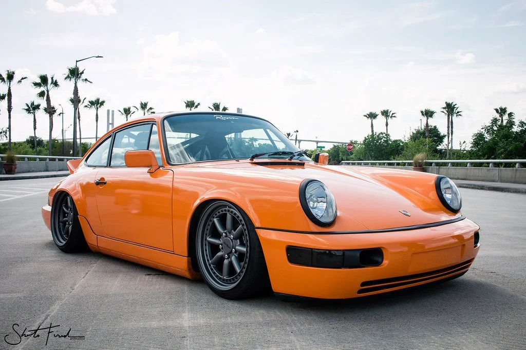 An orange classic Porsche 911 sports car parked on an outdoor concrete surface with a backdrop of palm trees and a cloudy sky.