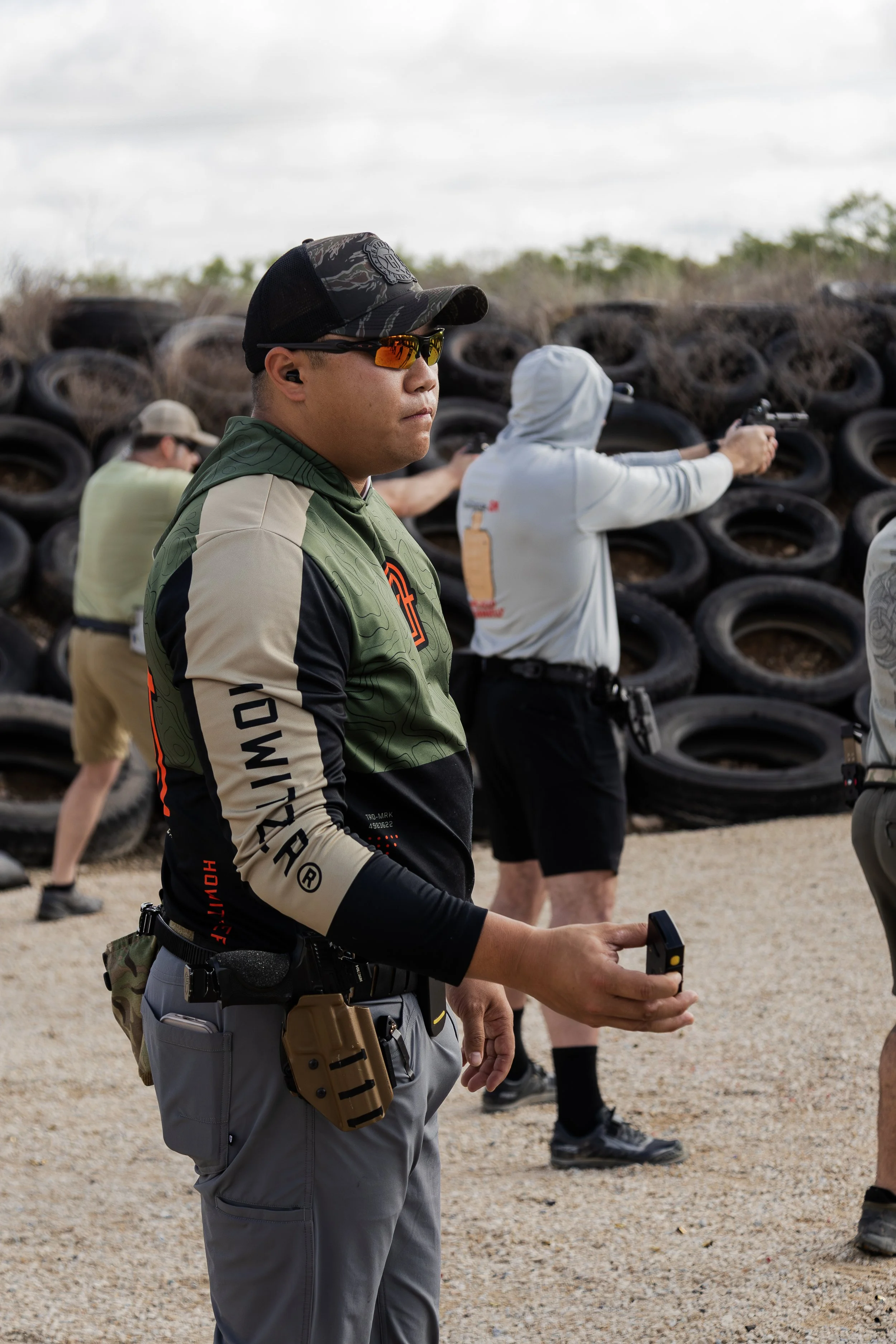 A man wearing sunglasses, a black and camouflage cap, ear protection, and tactical gear stands outdoors near a tire wall, holding a remote control, with other individuals shooting handguns in the background.